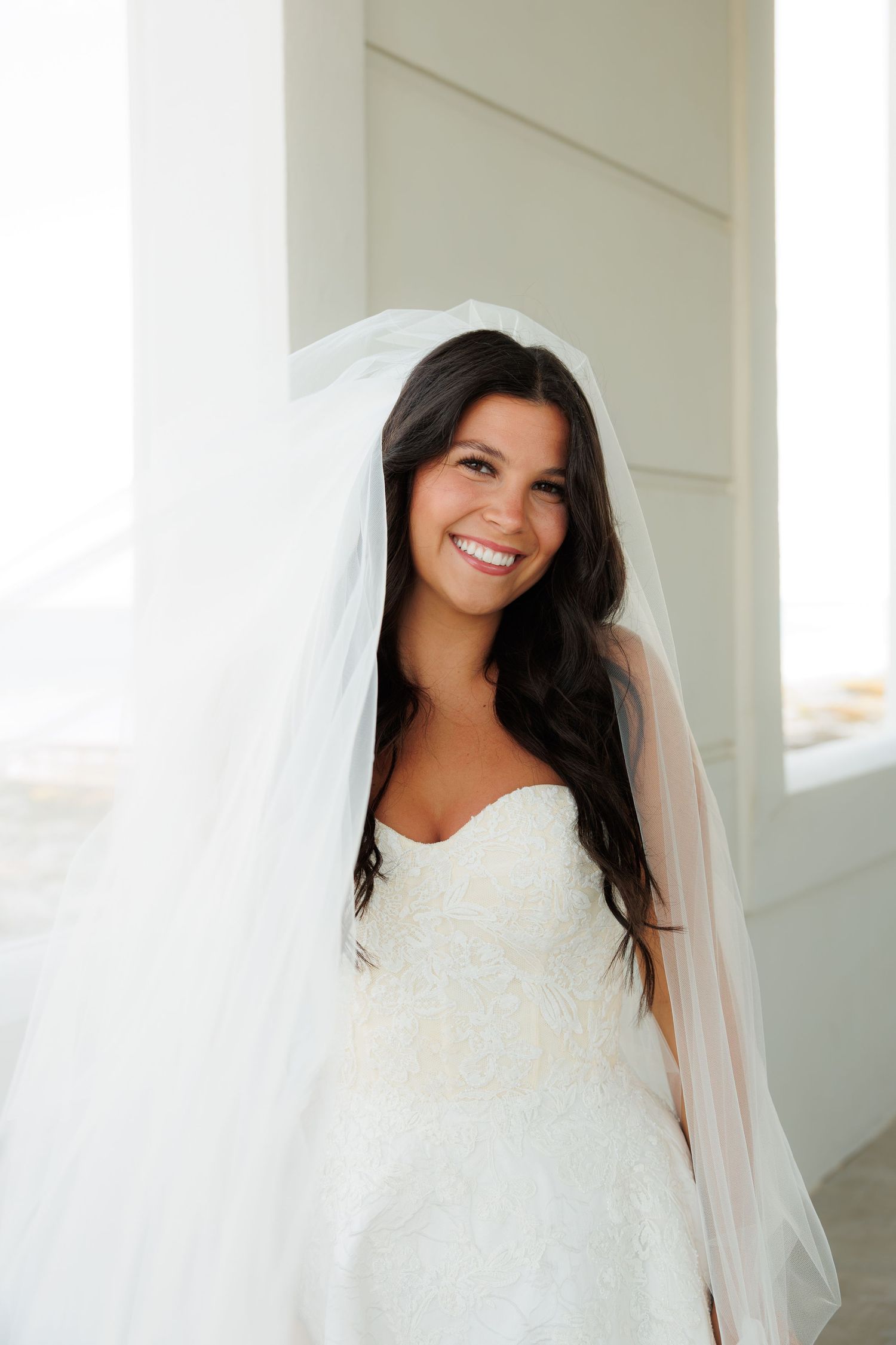 Smiling bride in white lace strapless gown and flowing veil poses by a column on her wedding day.