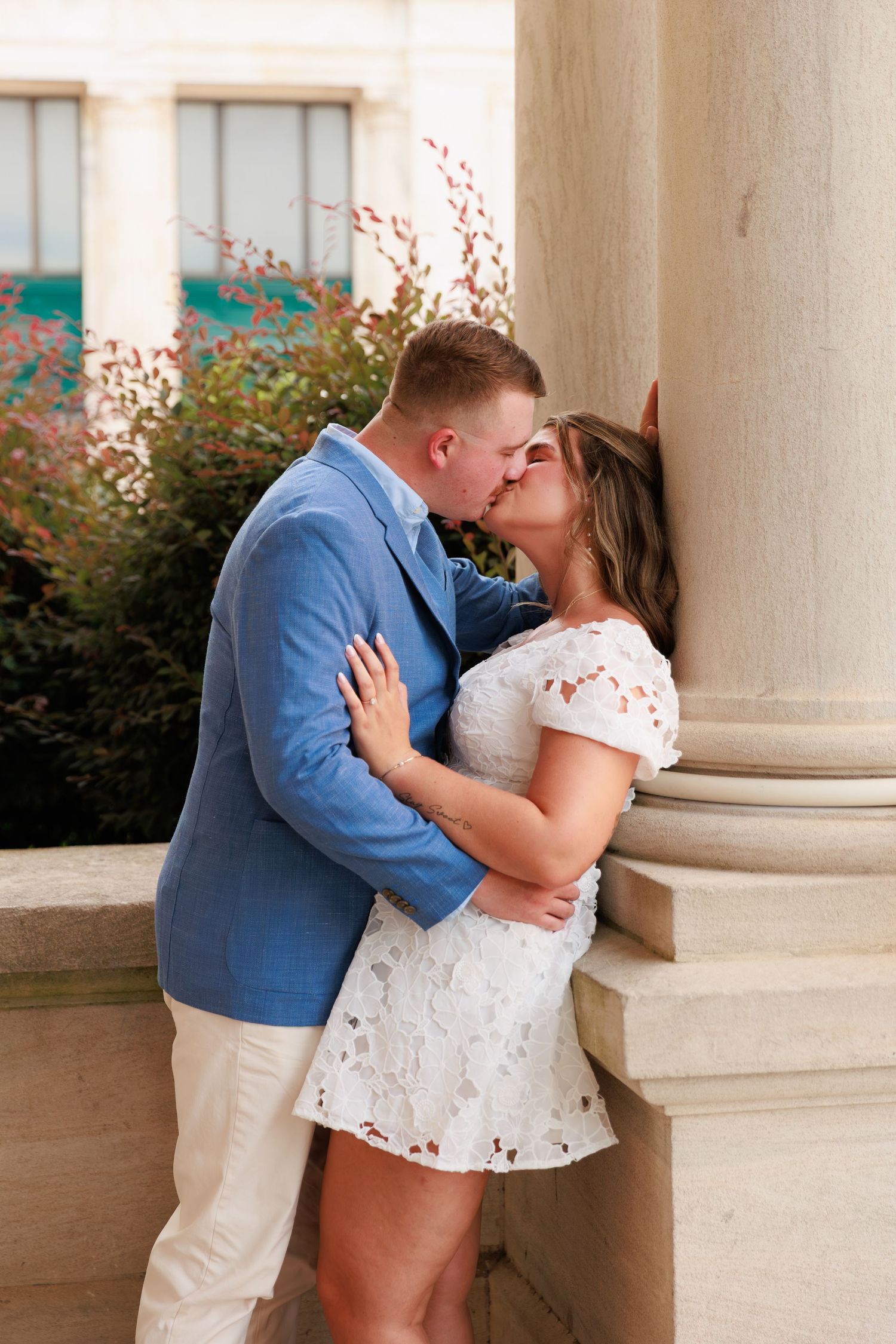 Couple sharing a passionate kiss against a stone column, man in blue blazer and woman in white lace mini dress.