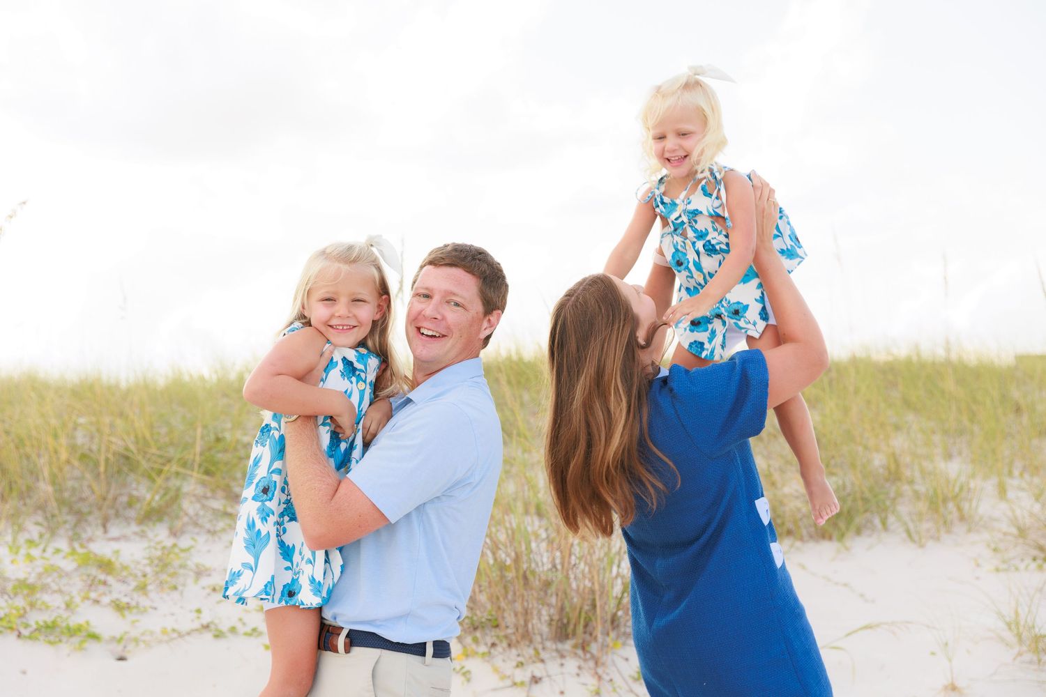 Family of four enjoying a sunny beach day, parents lifting two young girls in matching blue floral dresses.