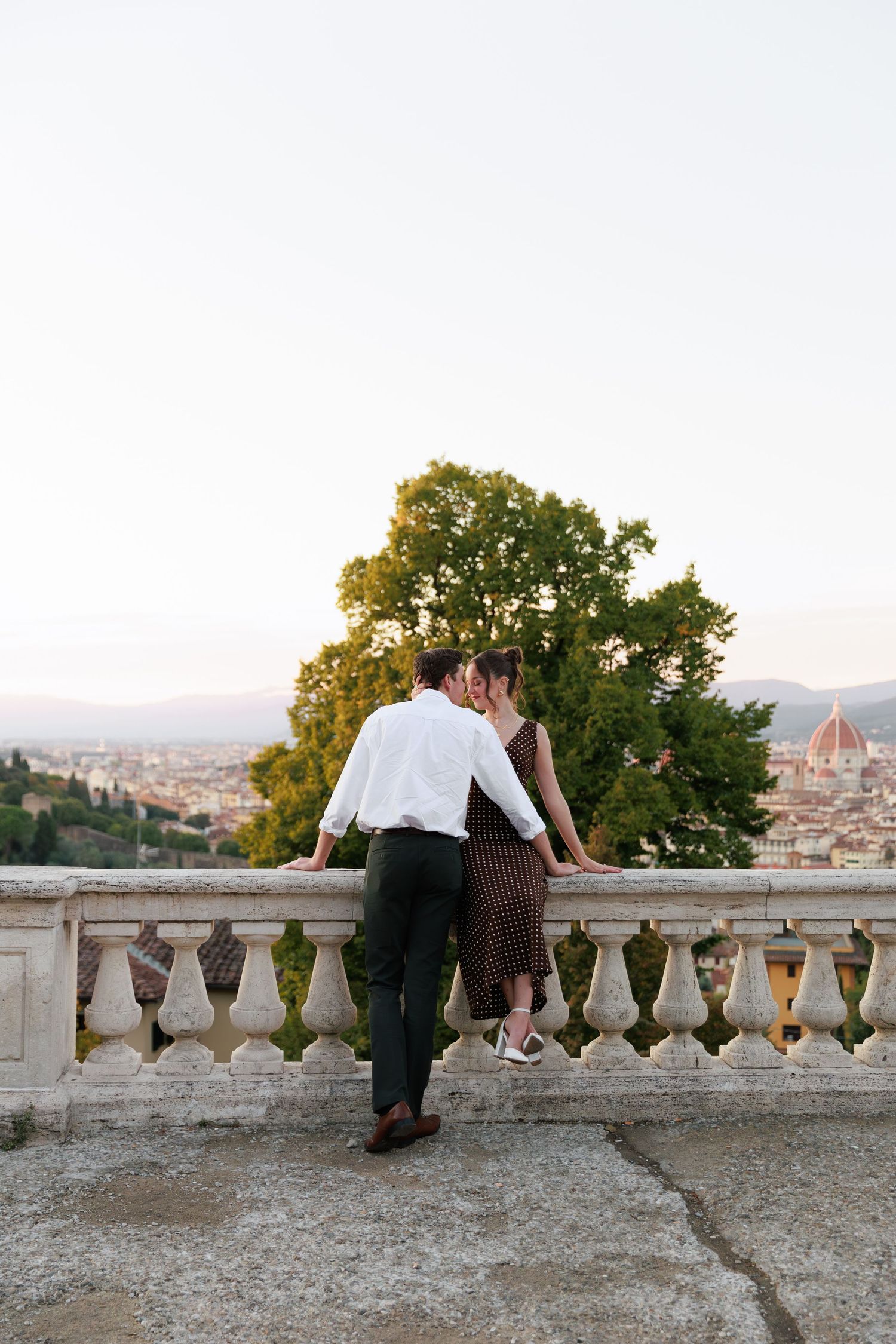 Couple sharing a romantic kiss on a stone balcony overlooking Florence skyline at sunset.