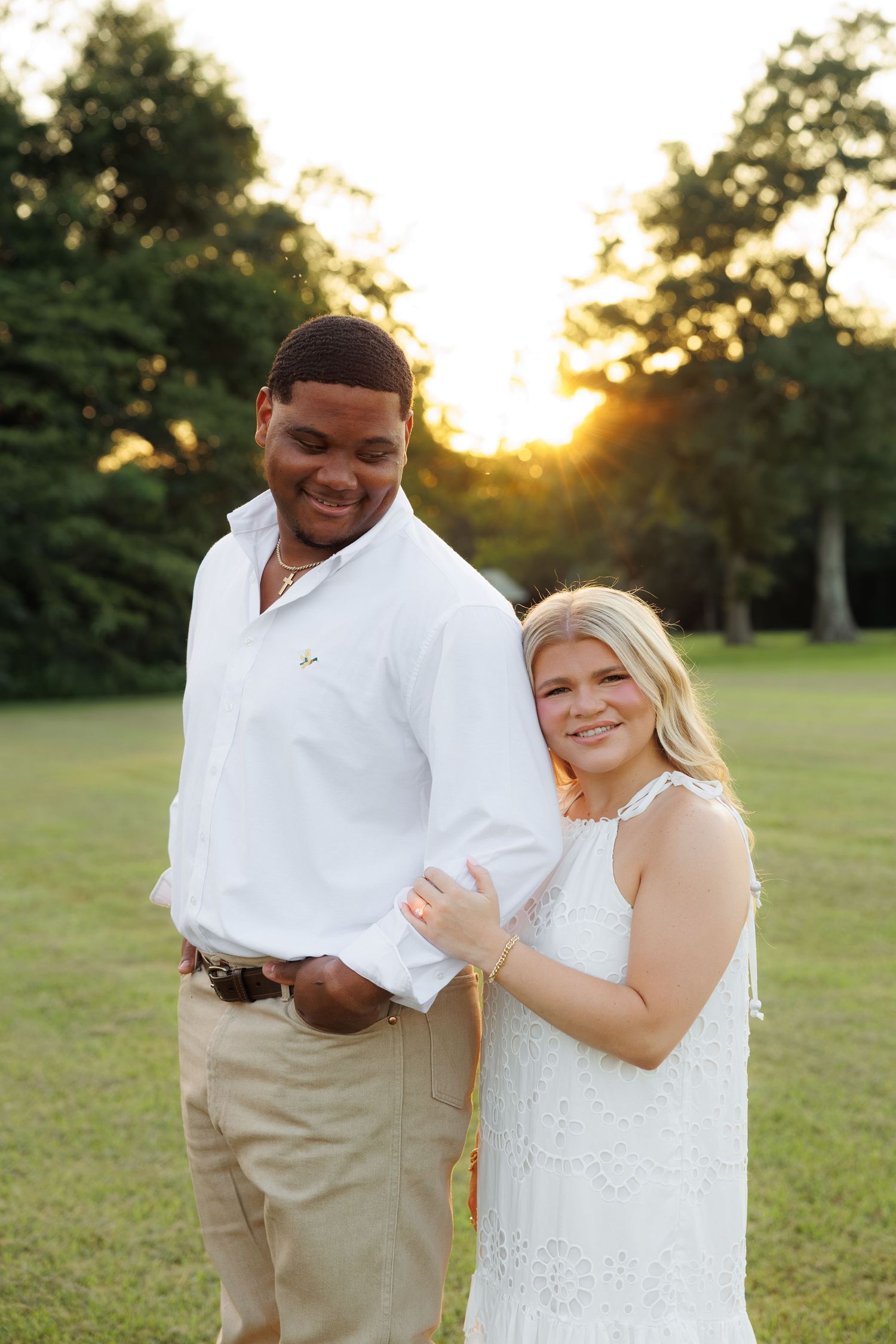 Couple in white outfits posing at golden hour in a lush green park, smiling warmly together.