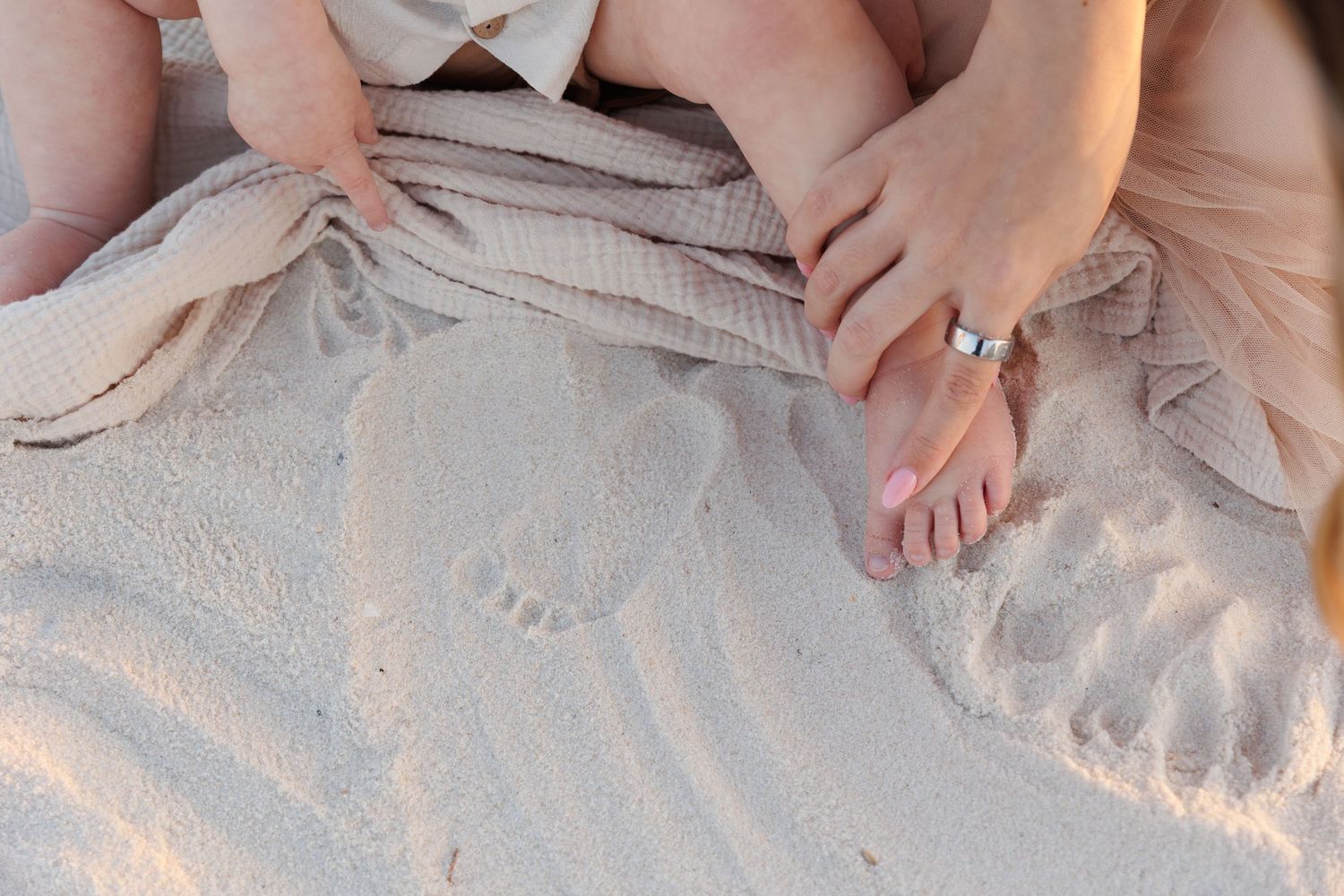 Adult and baby feet in sand at beach, tiny footprint visible, woman wearing silver ring on finger.