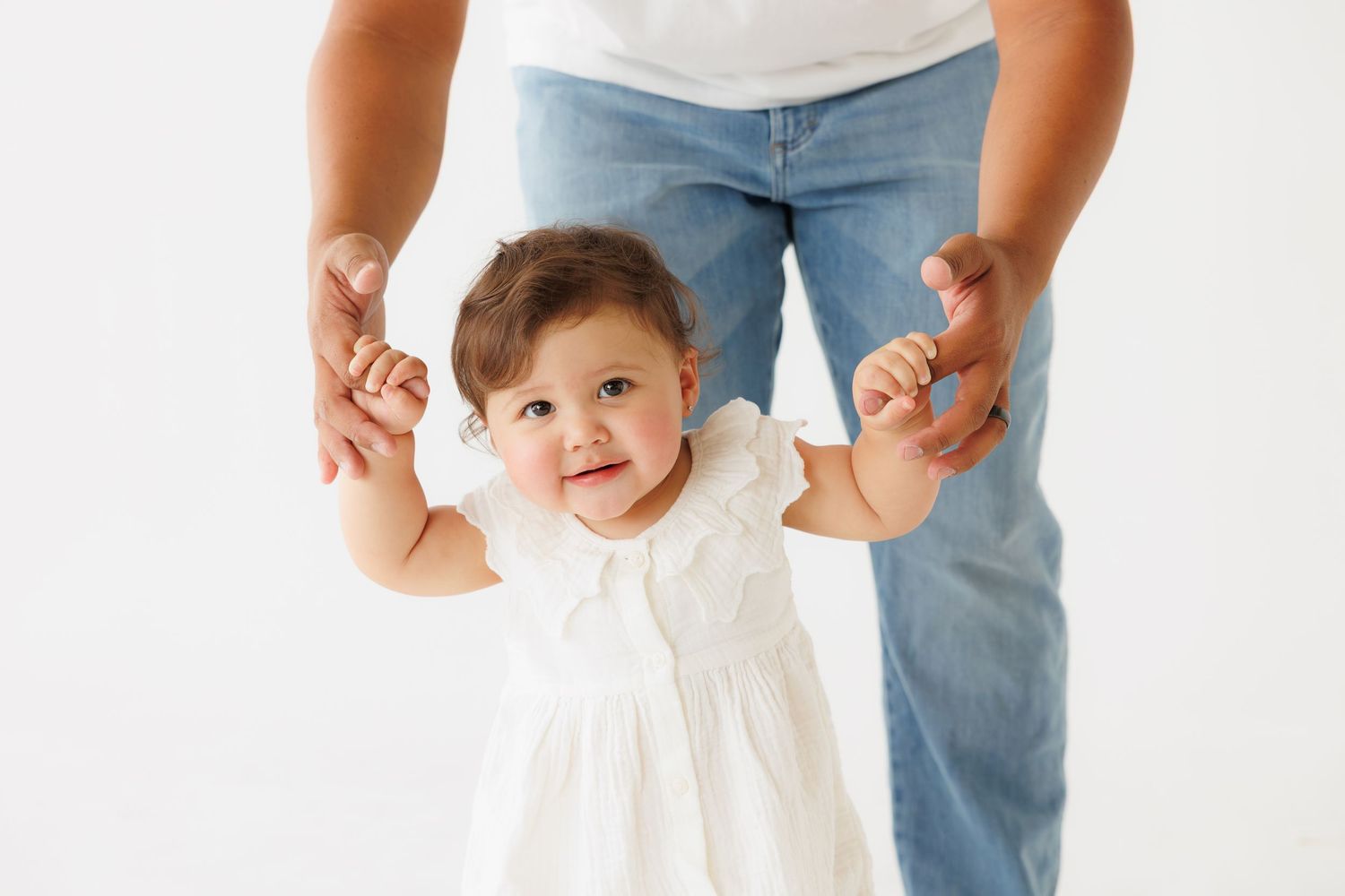 A baby girl in a white dress takes her first steps while being supported by an adult holding her hands.
