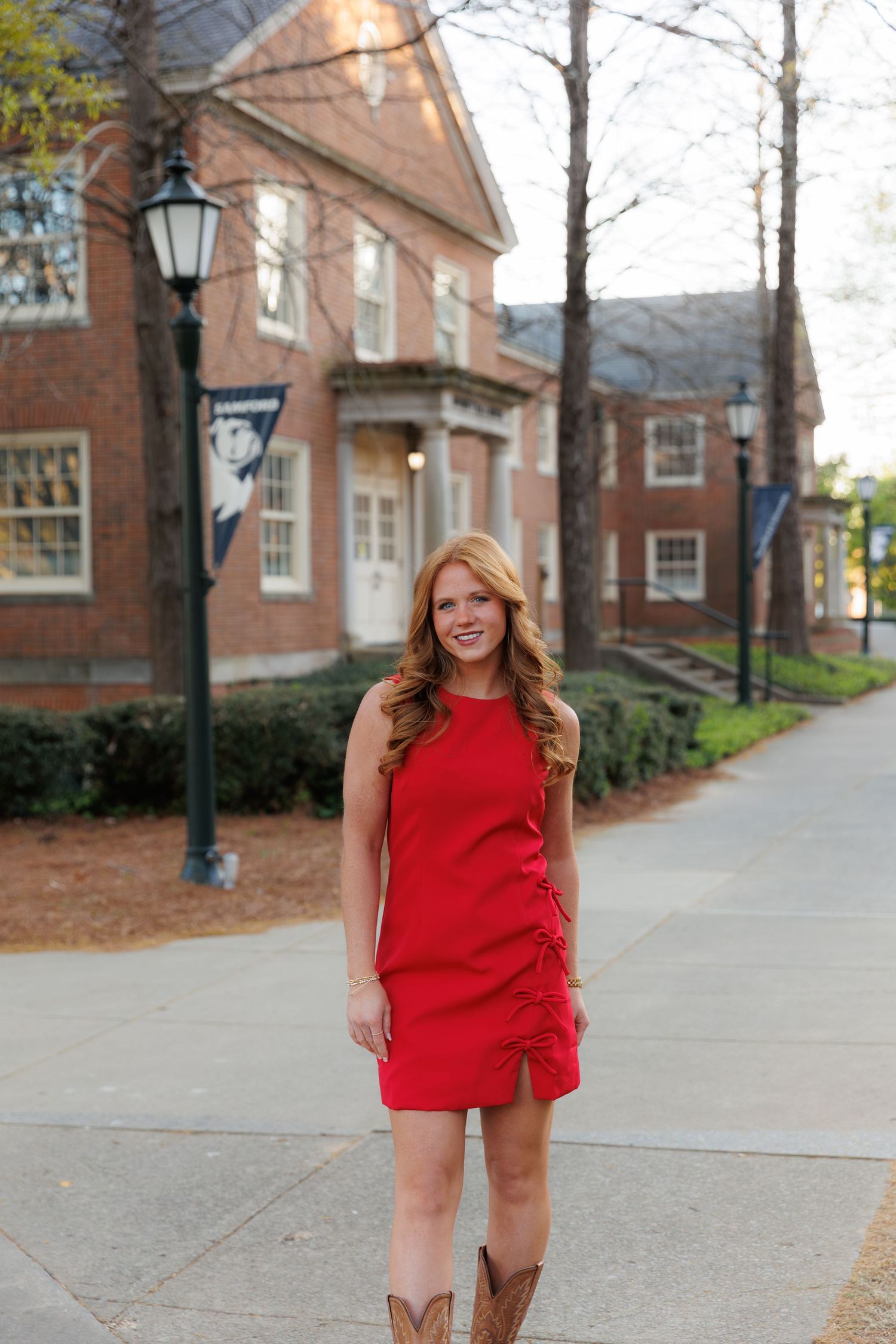 Young woman in red sleeveless dress and cowboy boots smiling on a brick campus walkway near a lamppost.