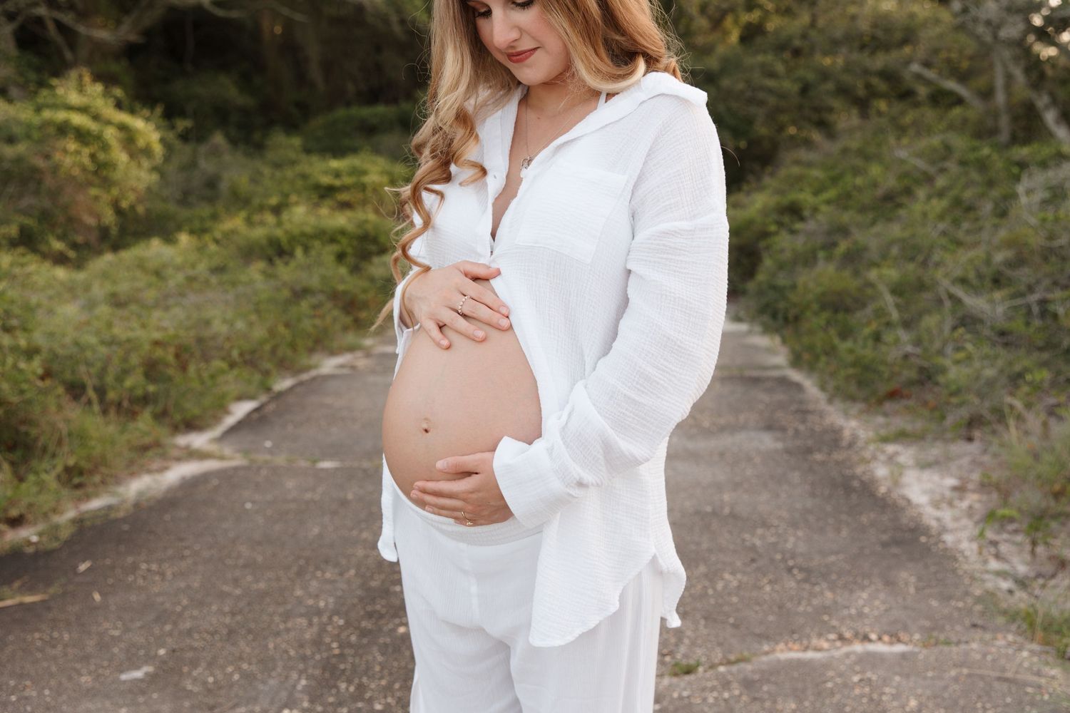 A person in a white dress cradles their pregnant belly while standing on a forest path during a maternity photoshoot.