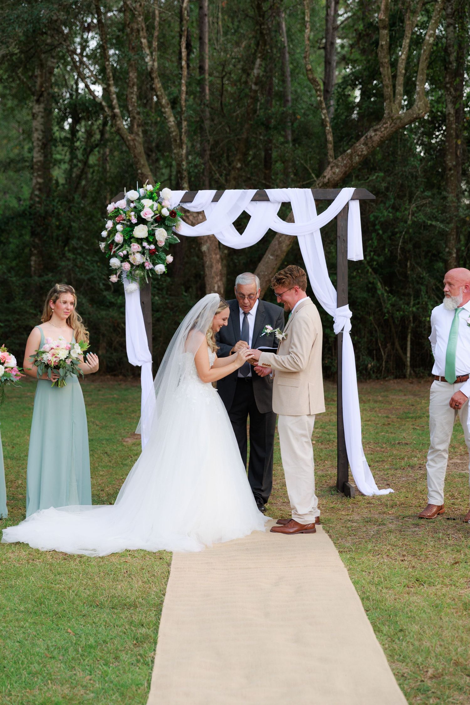 Outdoor wedding ceremony under trees with white draped arch and mint green bridesmaids dresses.