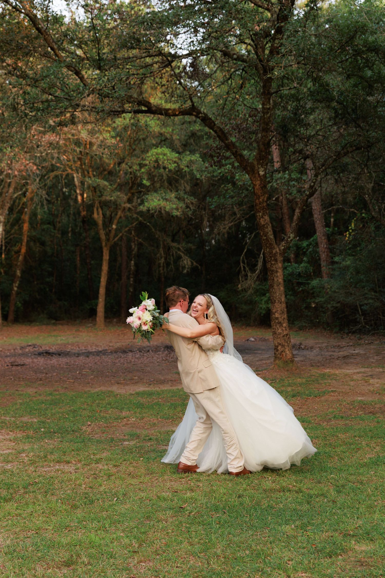 A romantic wedding couple embraces on the grass in a serene wooded setting during their outdoor photoshoot.