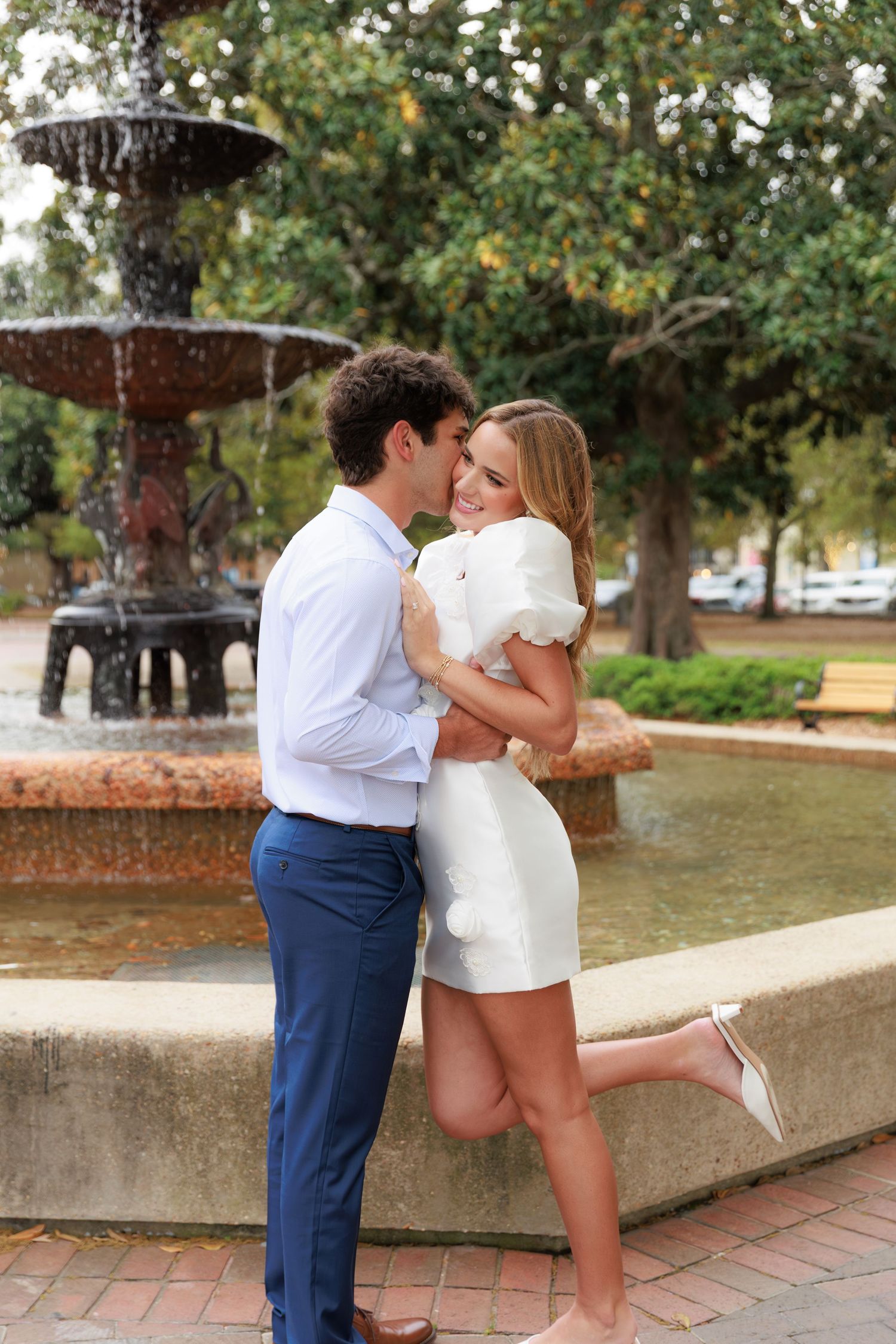 A romantic moment shared between a couple by a fountain, with her wearing a white dress and him in blue slacks.
