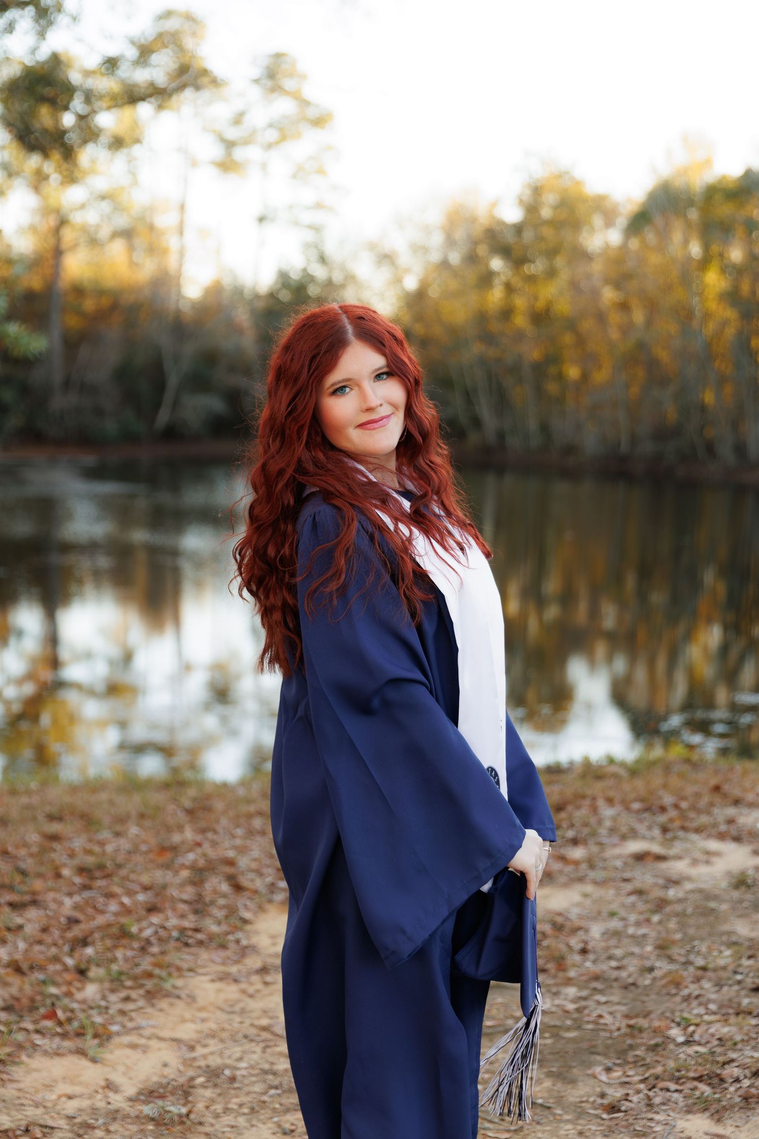 Woman with red hair poses by a lake in autumn wearing a navy blazer and white top.