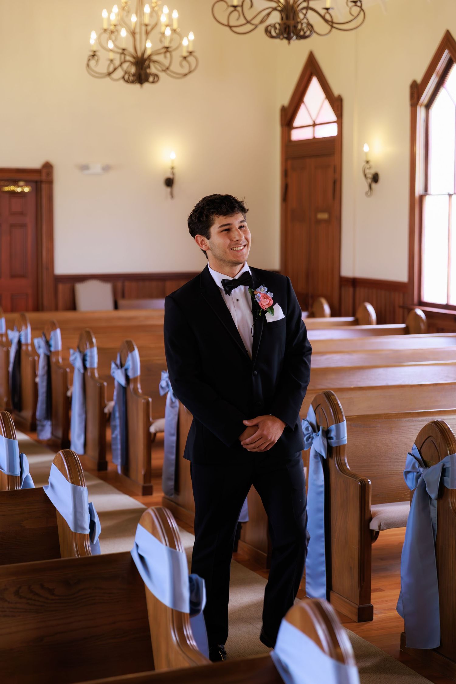 A person in a black suit and red boutonniere poses in a church sanctuary decorated with blue bows on wooden pews.