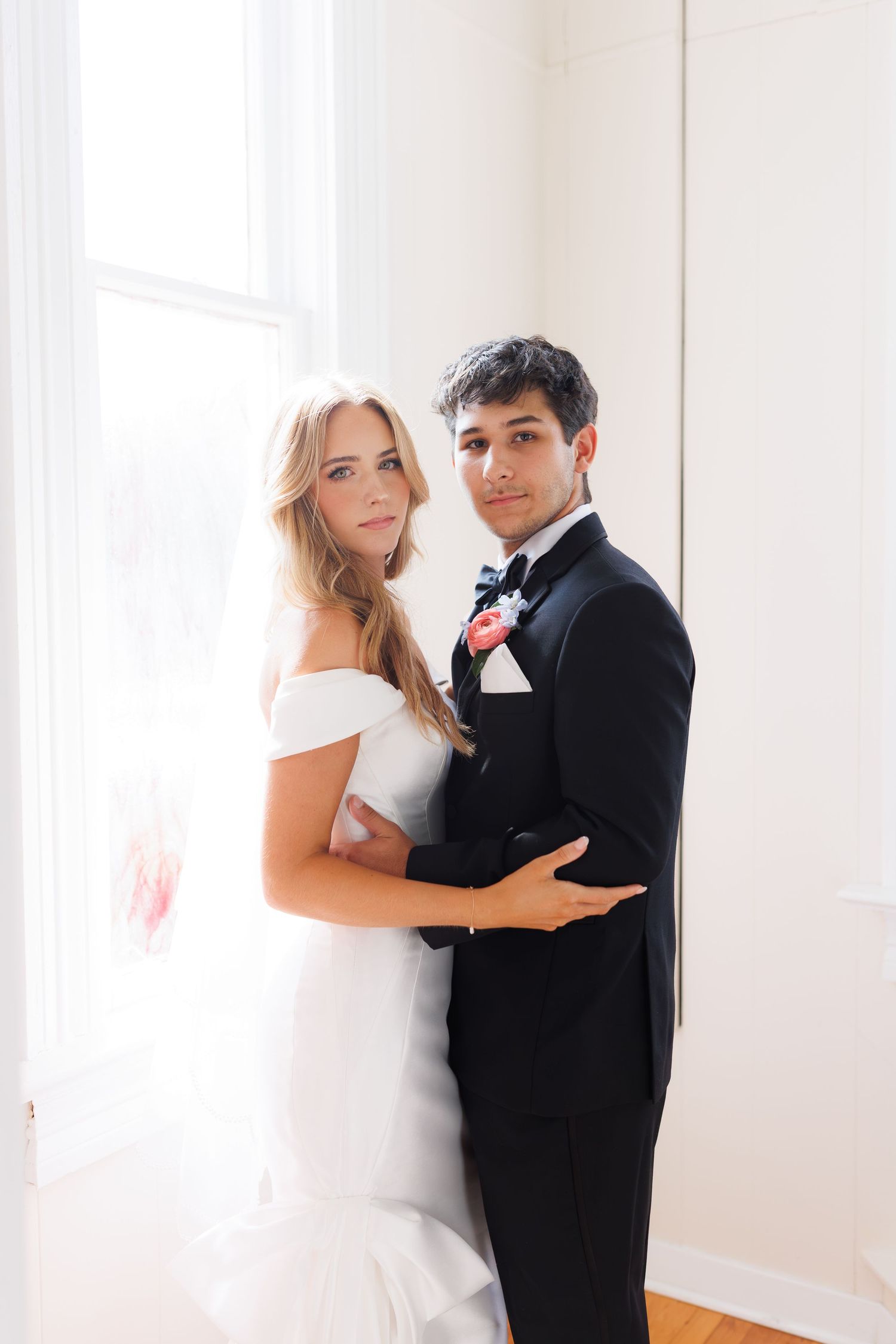 A romantic indoor wedding portrait of a couple in formal wedding attire standing by a bright window.