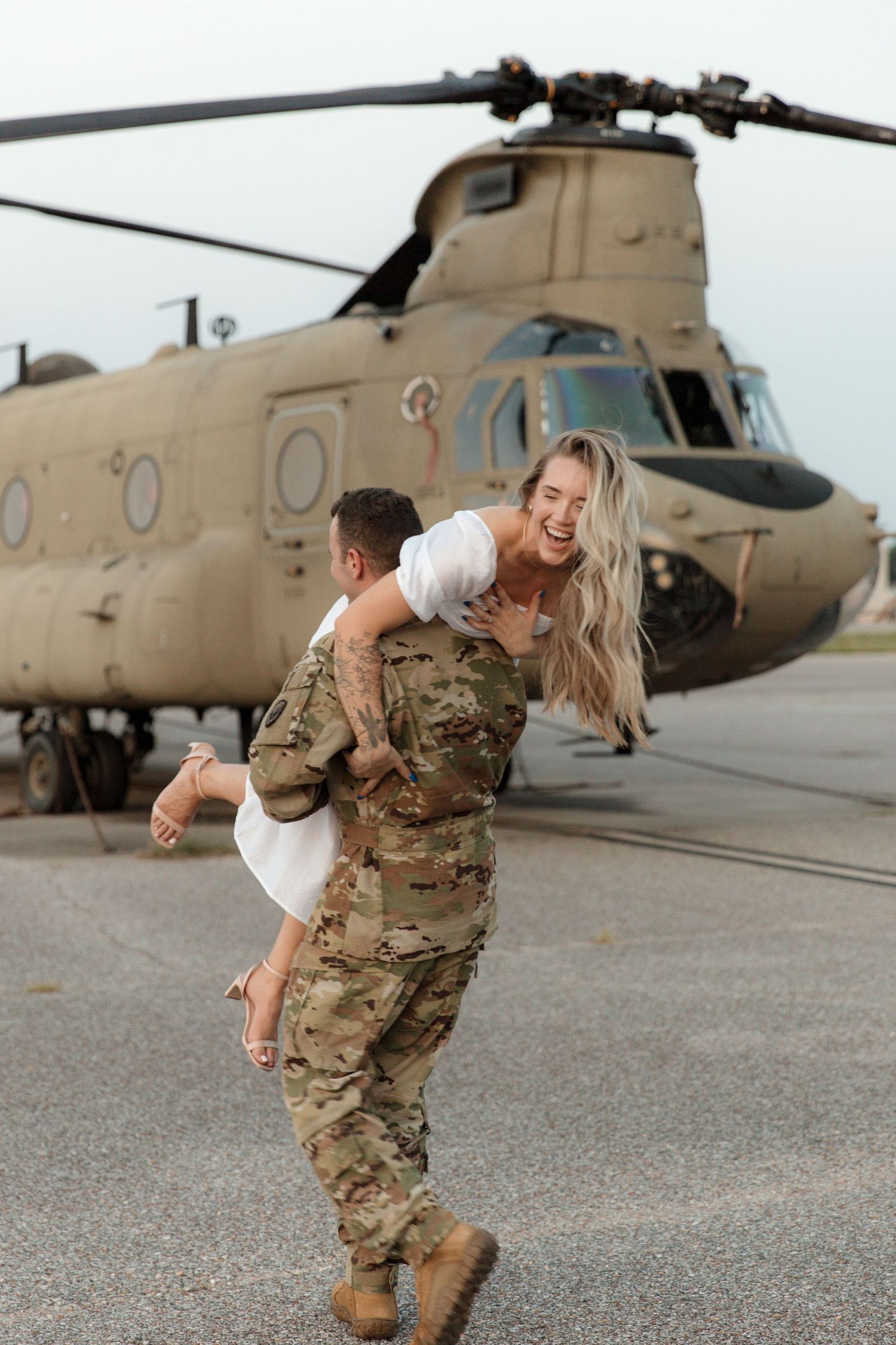 Service member carries partner playfully in front of military helicopter.