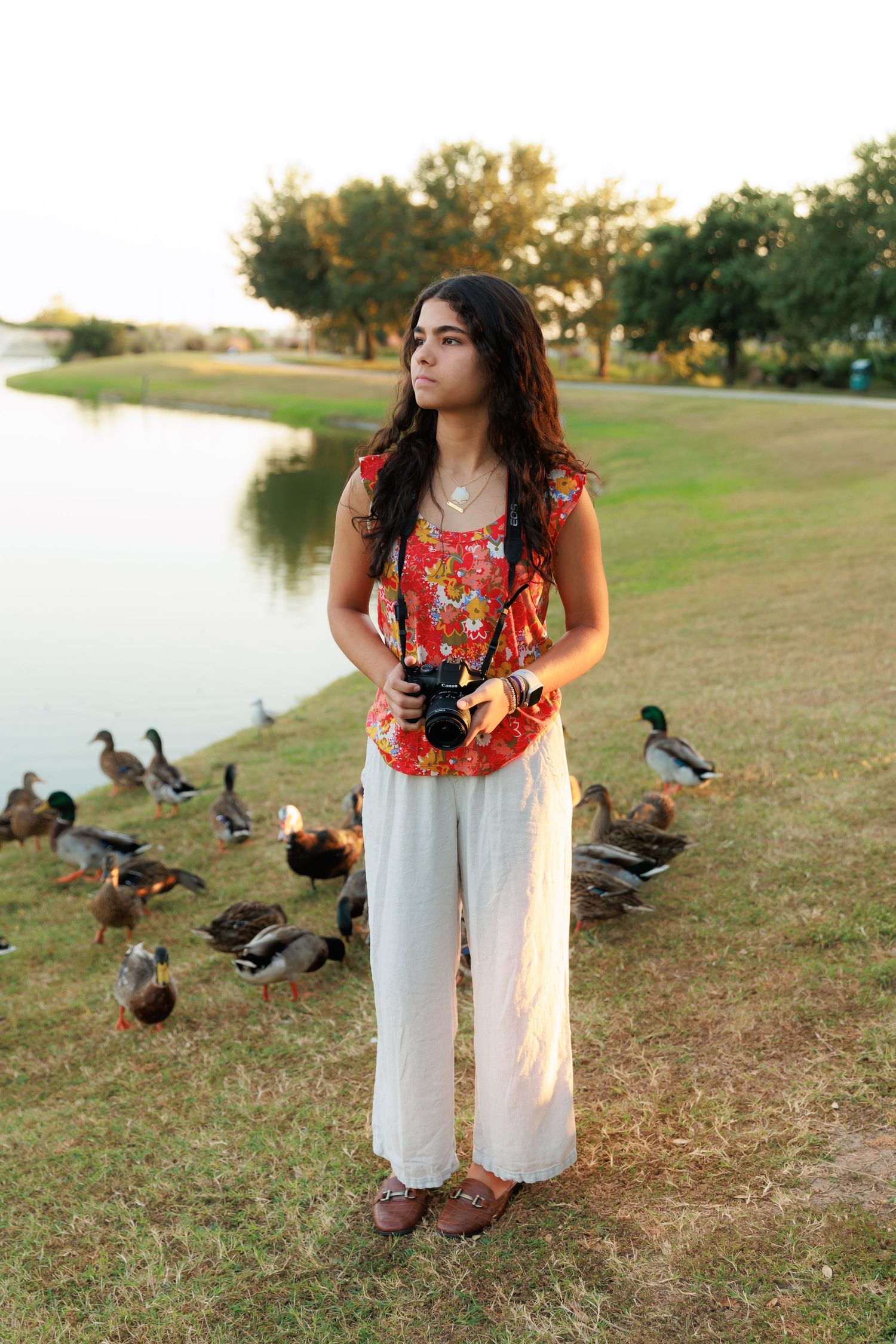Photographer in floral red top and white pants stands by a lake with ducks during golden hour.