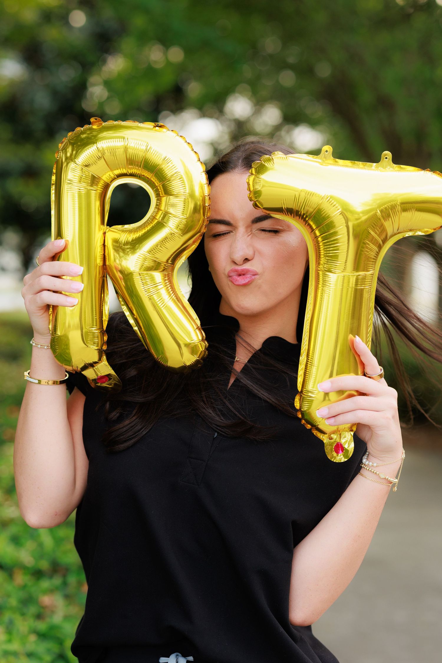 Young woman holding gold letter R and T balloons outdoors, making a kissing face, wearing a black top.