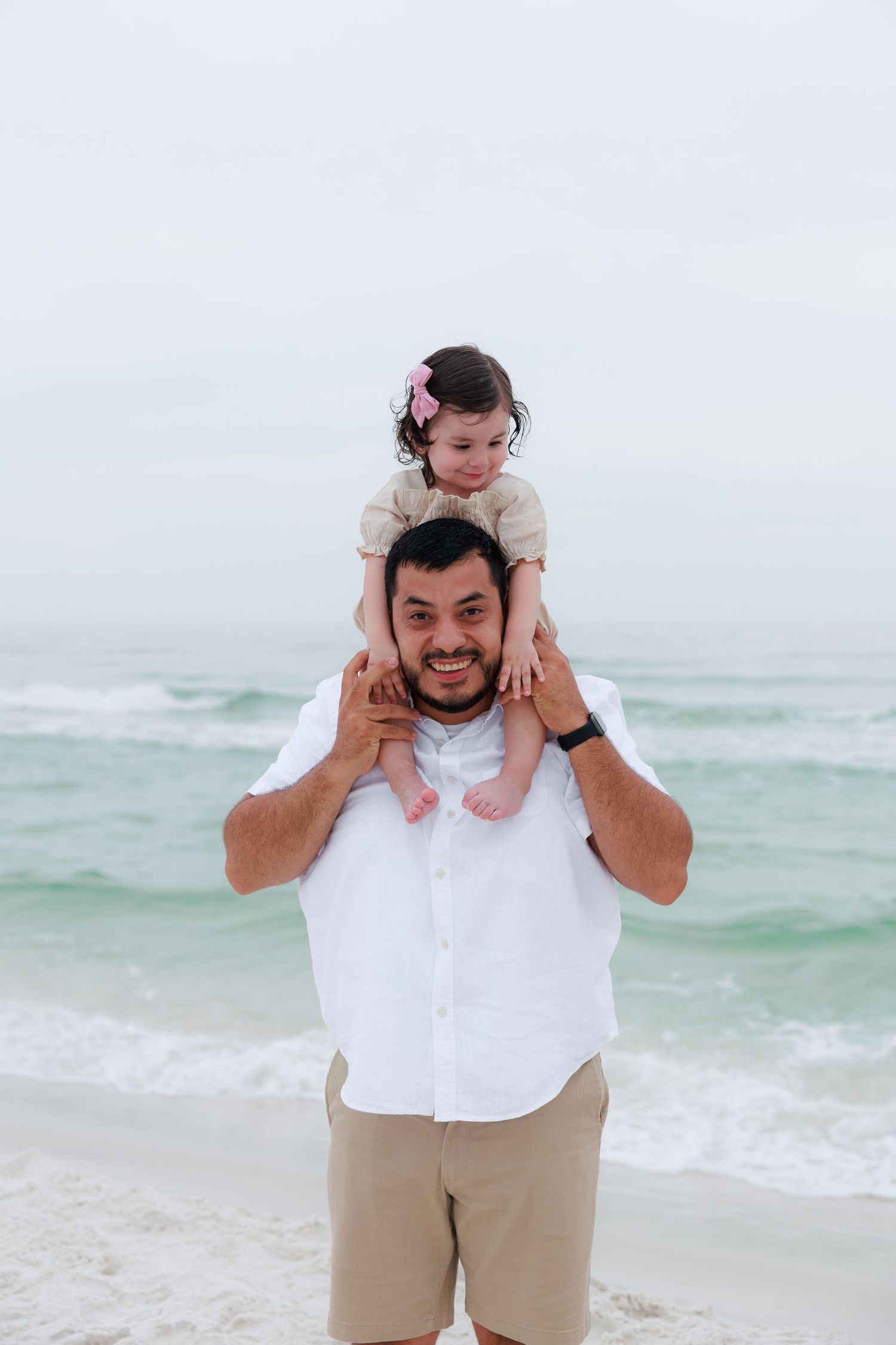 A smiling man carries a baby girl on his shoulders at a beach with turquoise waves behind them.