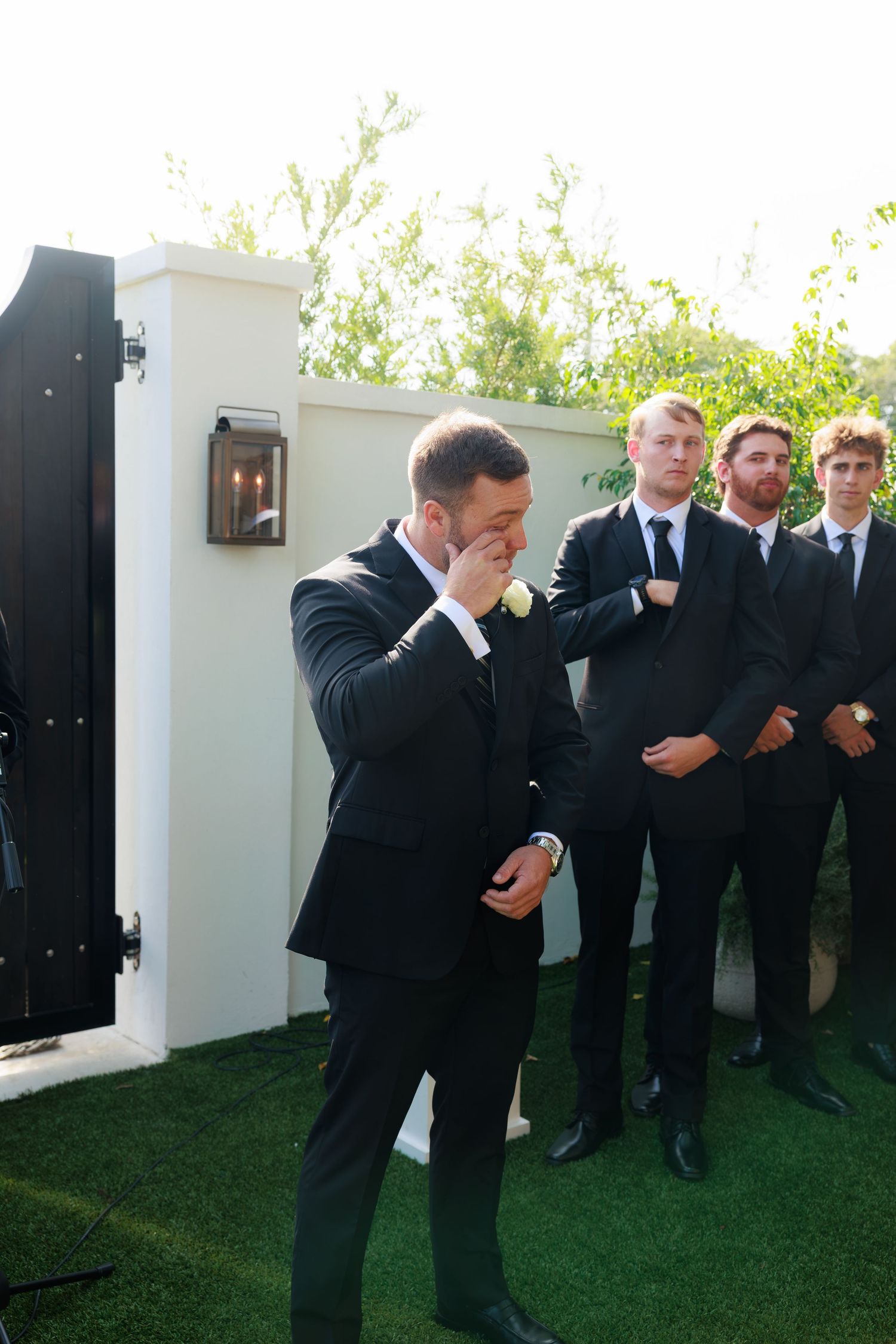 Emotional groom crying before wedding ceremony, standing outdoors with groomsmen in black suits behind him.
