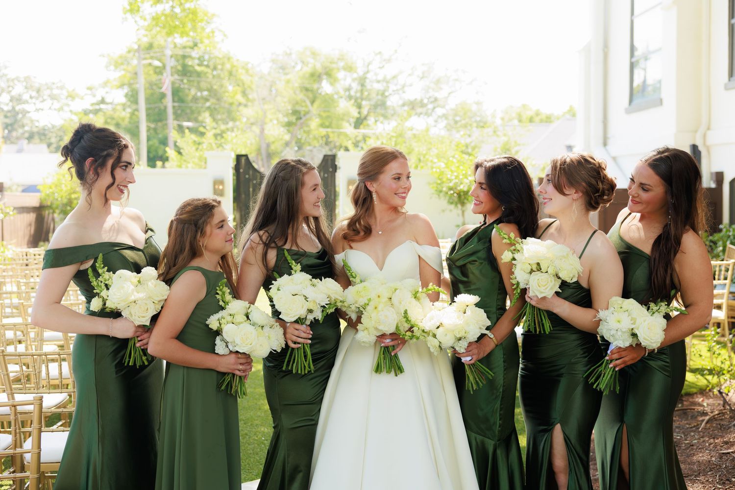 Bride in white gown with bridesmaids in emerald green dresses holding white bouquets at outdoor wedding ceremony.