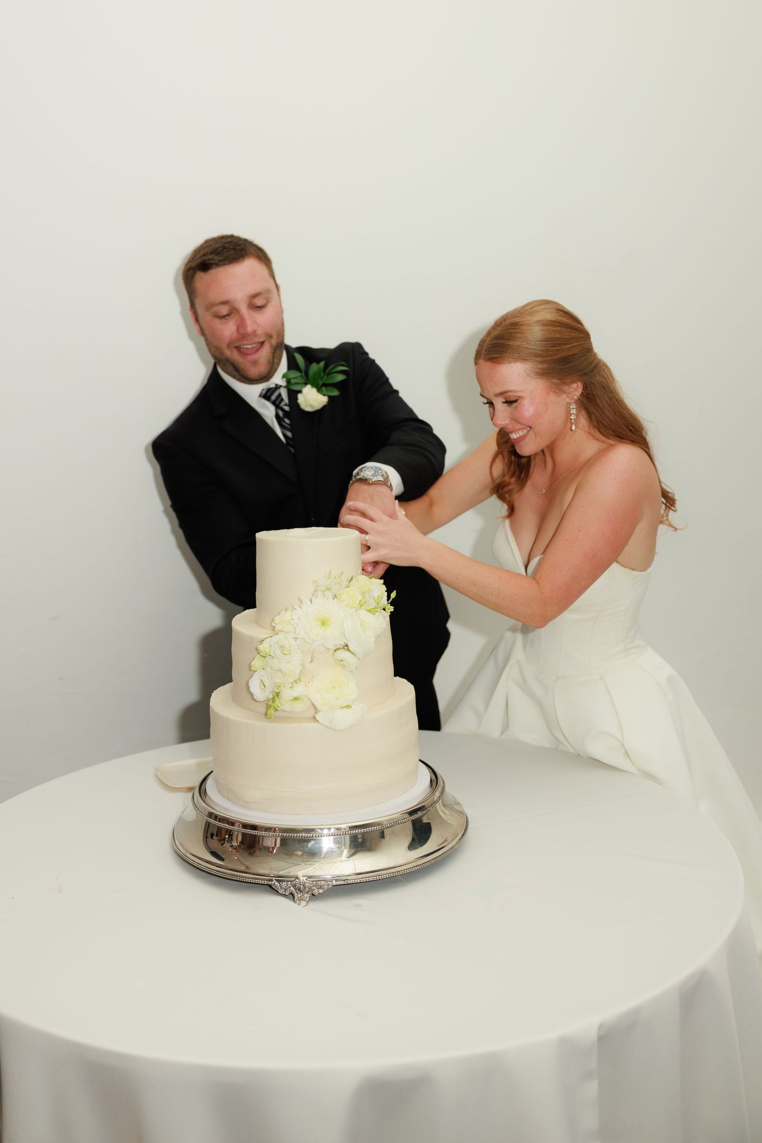 Bride and groom laugh together while cutting elegant three-tier white wedding cake decorated with flowers.