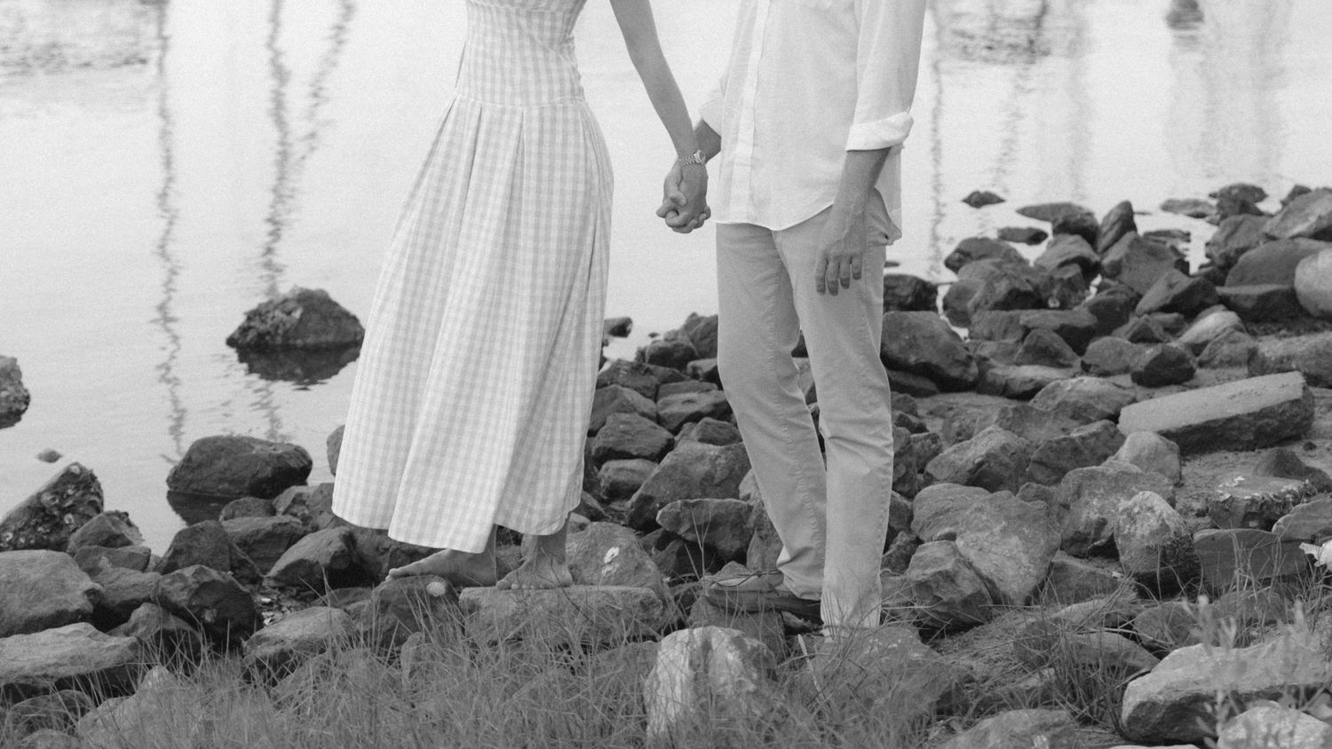 A couple holds hands while standing on rocky shoreline near water, wearing casual summer outfits in a black and white photo.