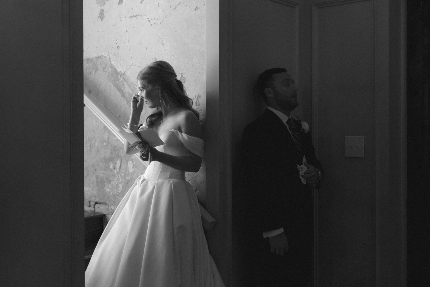 Black and white photo of bride in strapless gown and groom in suit standing back-to-back in doorway.
