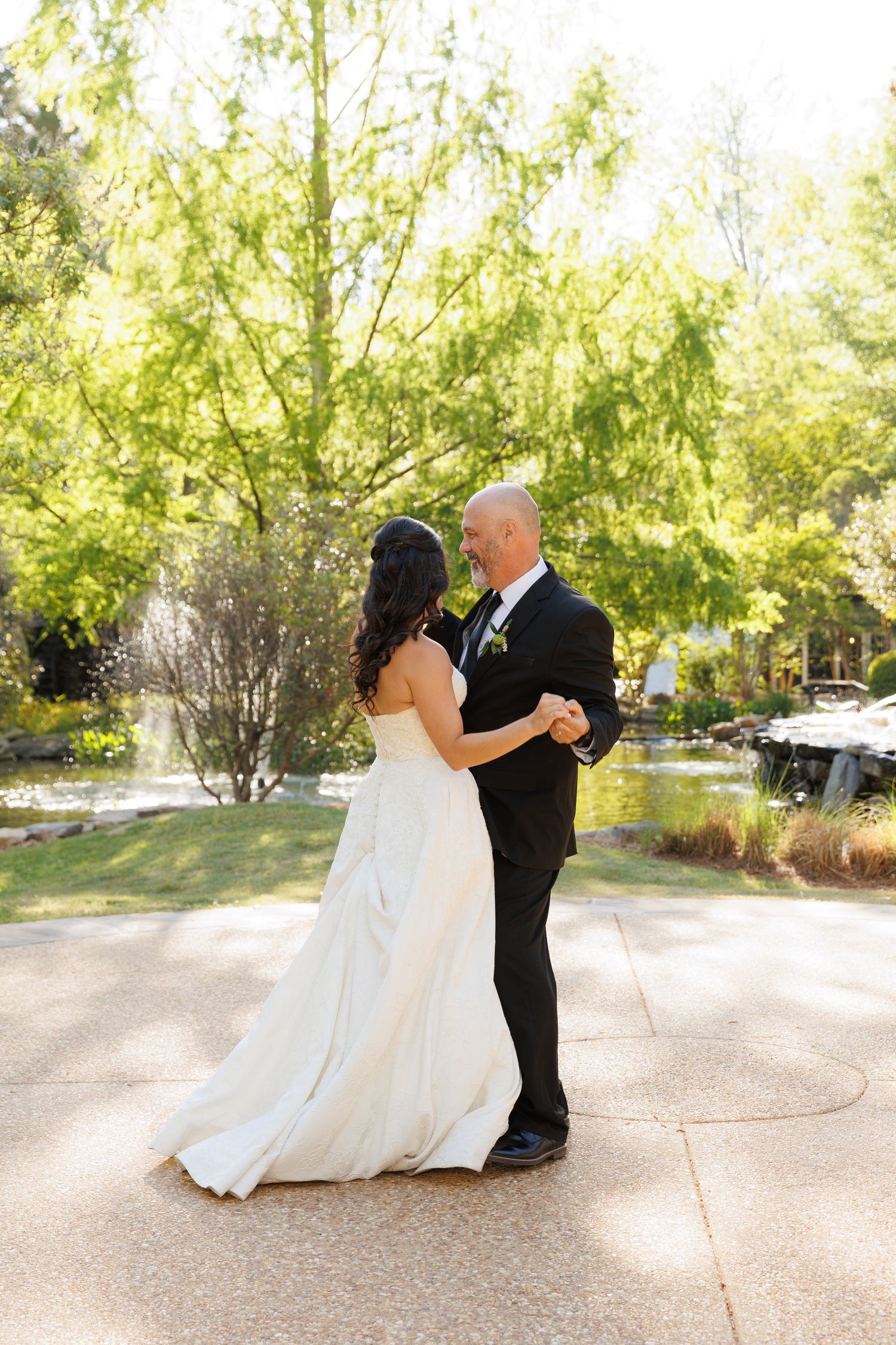Smiling bride and groom share a first dance outdoors in a bright, green garden near a serene pond.