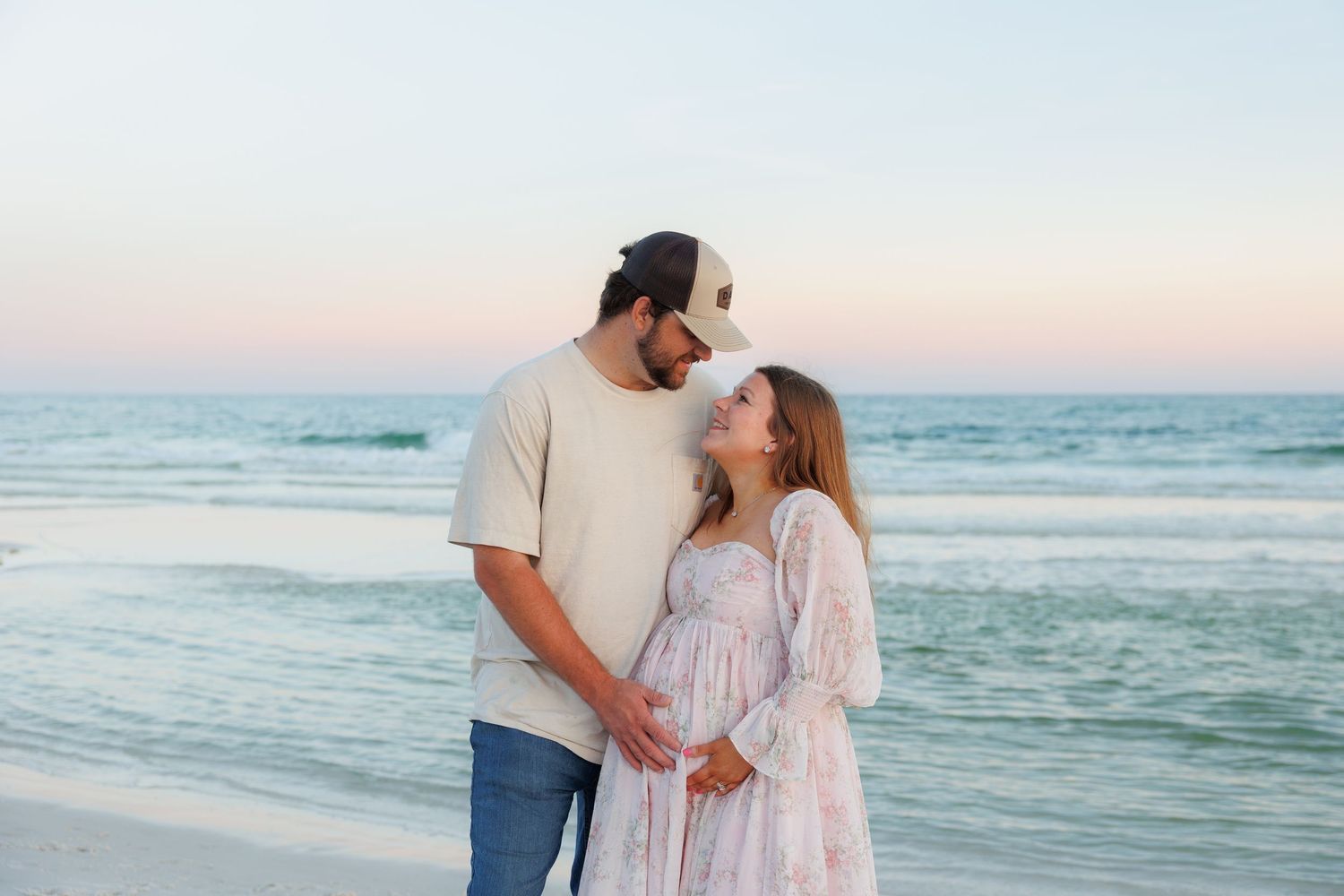 Couple sharing a tender moment on the beach at sunset, woman in floral maternity dress cradling baby bump.