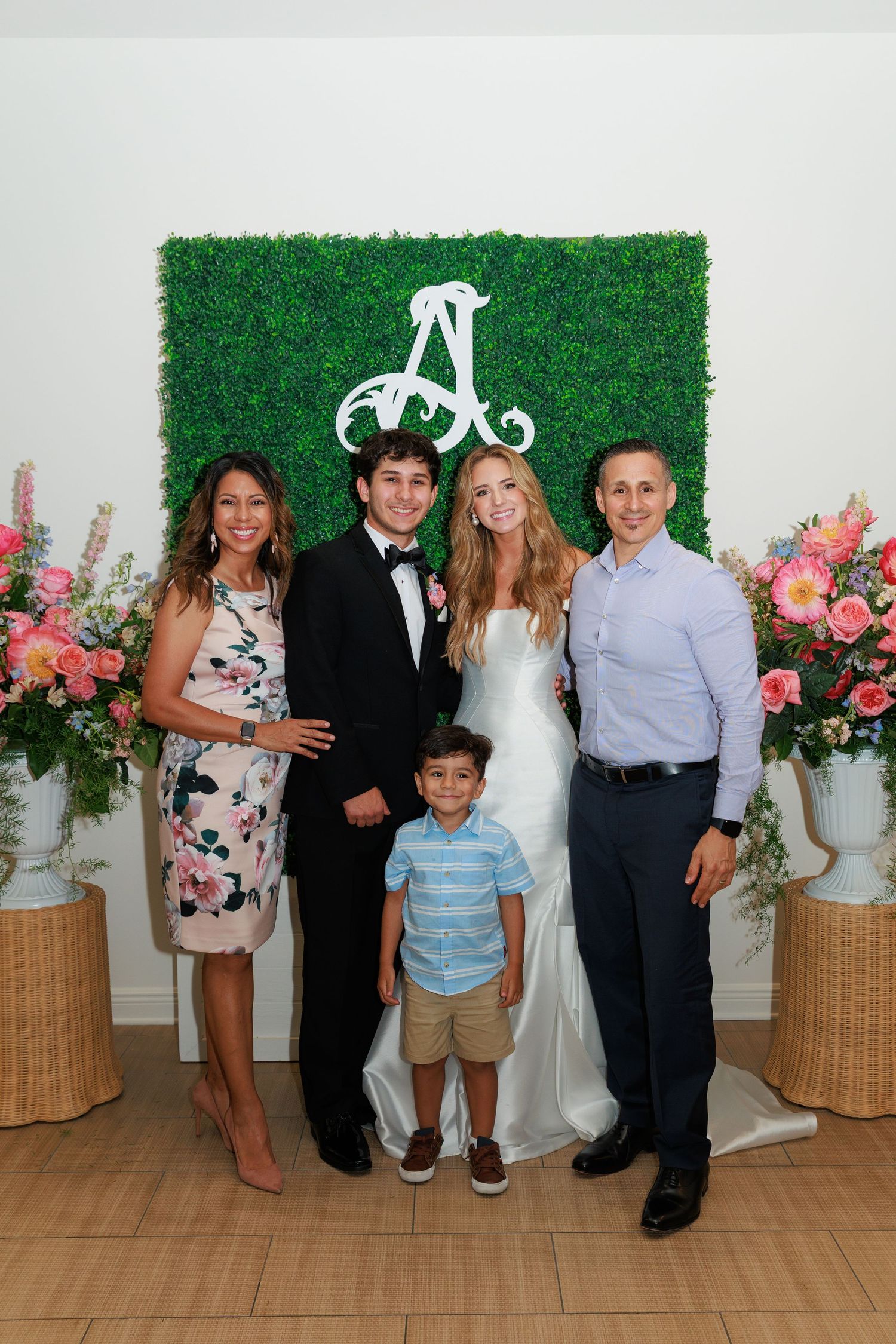 Group photo at wedding reception with green hedge wall backdrop, floral arrangements, and decorative monogram letter A above.