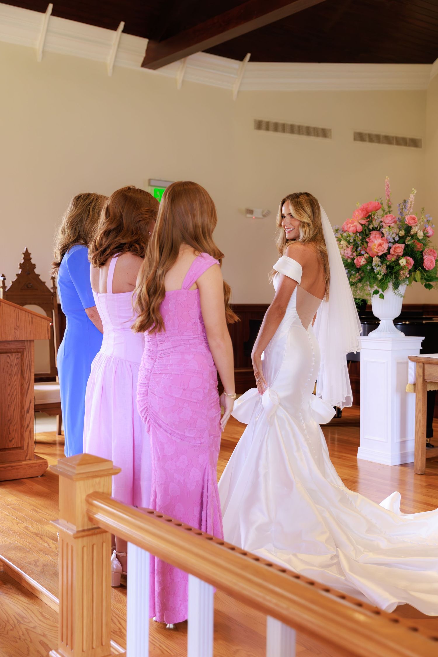 Inside a wedding chapel with bridesmaids in pink and blue dresses gathered near a bride in a white gown and veil.