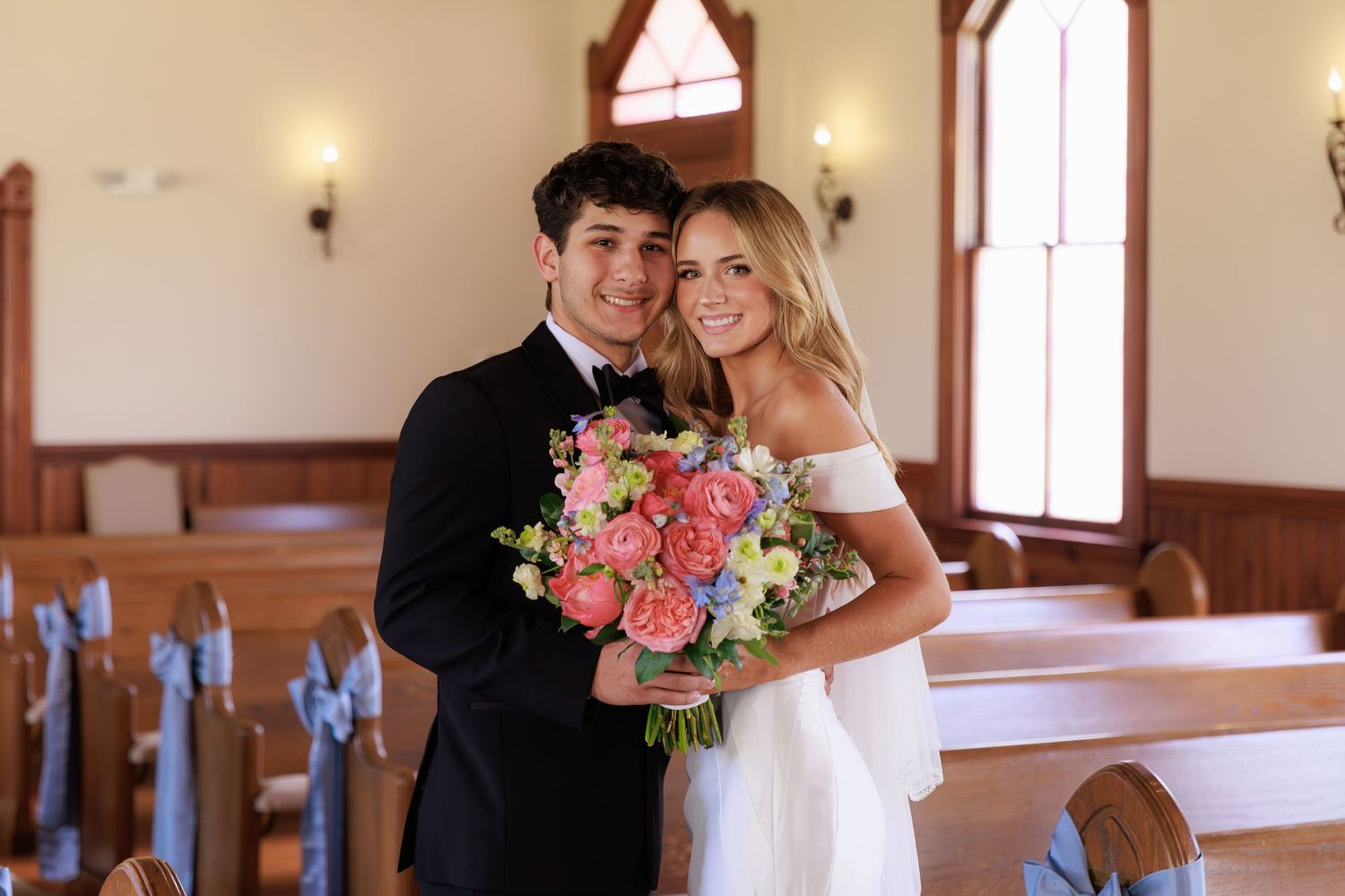 A wedding couple stands together in a chapel holding a beautiful pink and white flower bouquet.