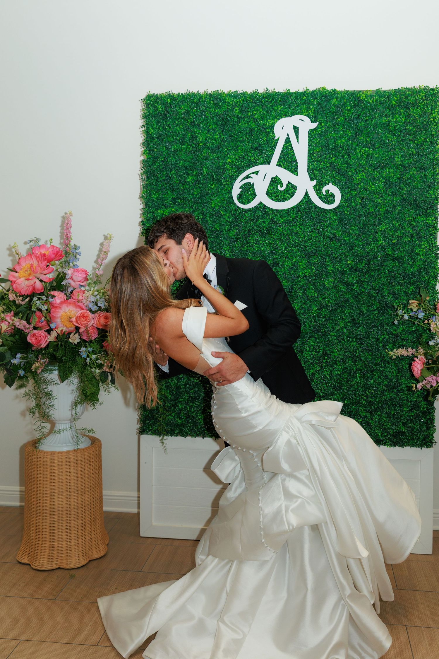 A romantic dance dip with wedding attire against a green hedge wall backdrop with pink floral arrangements.