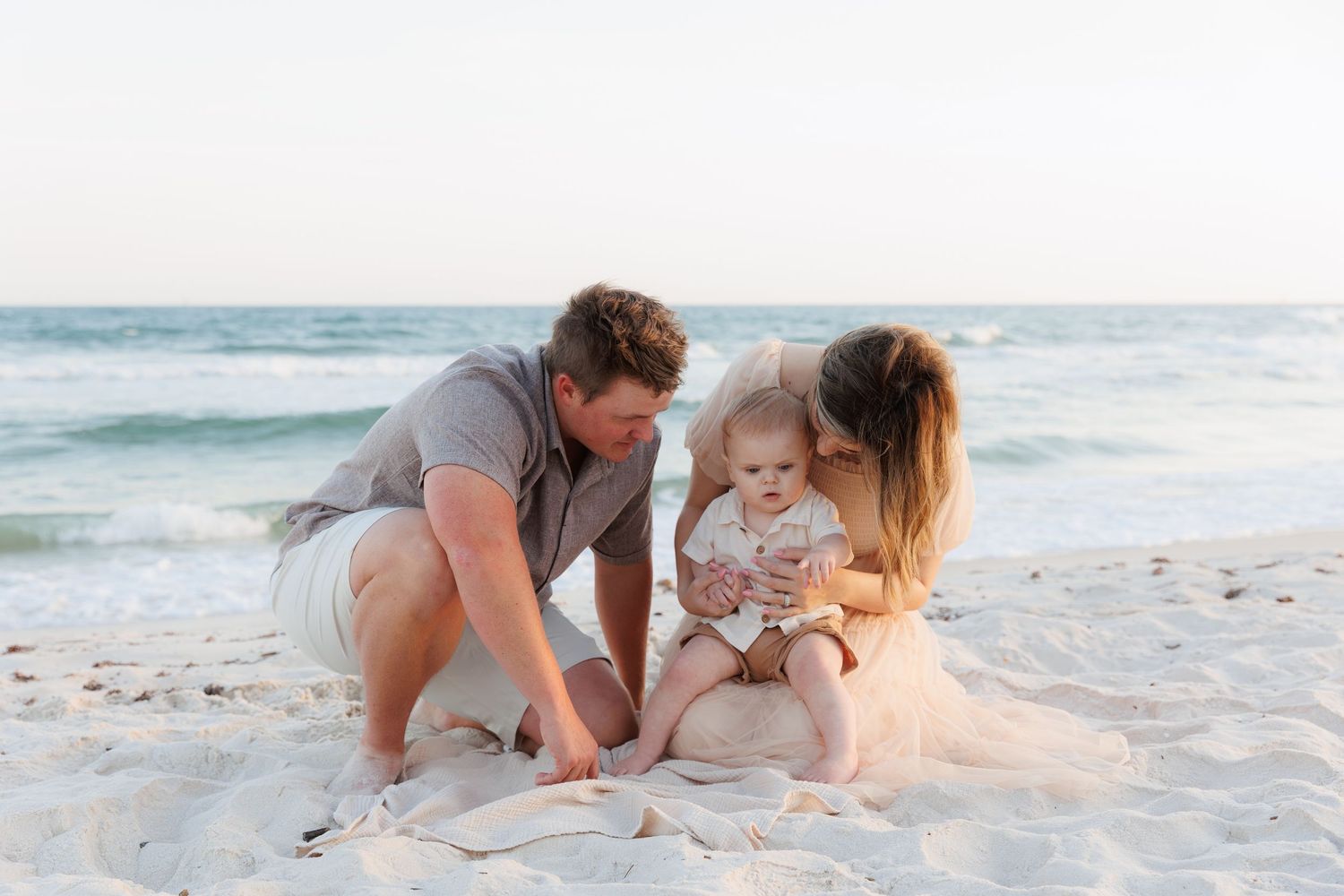 A young family with a baby sits on a sandy beach, parents lovingly kissing their child near the ocean.