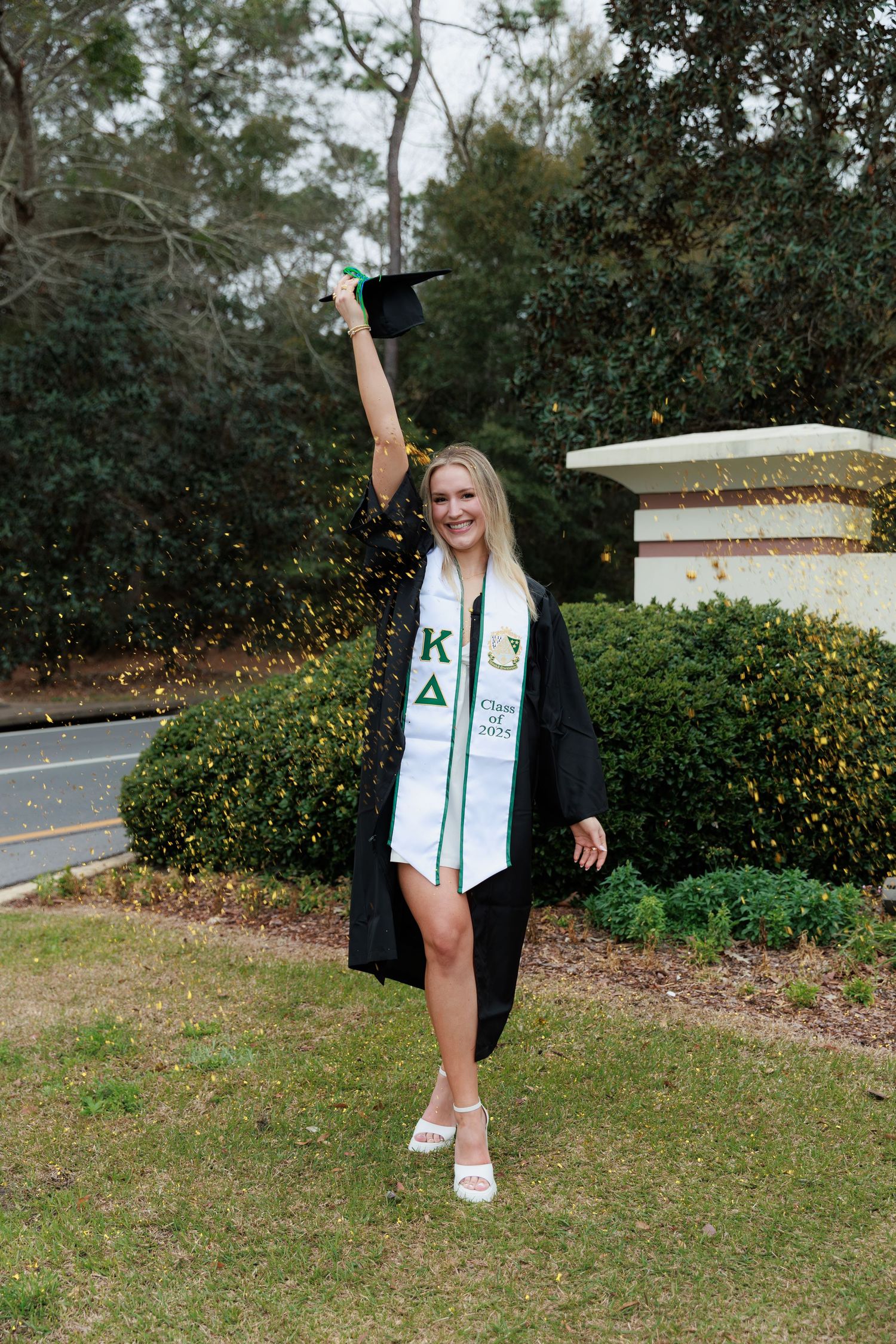 Blonde graduate in black gown and Kappa Delta stole joyfully raising cap near campus entrance sign.