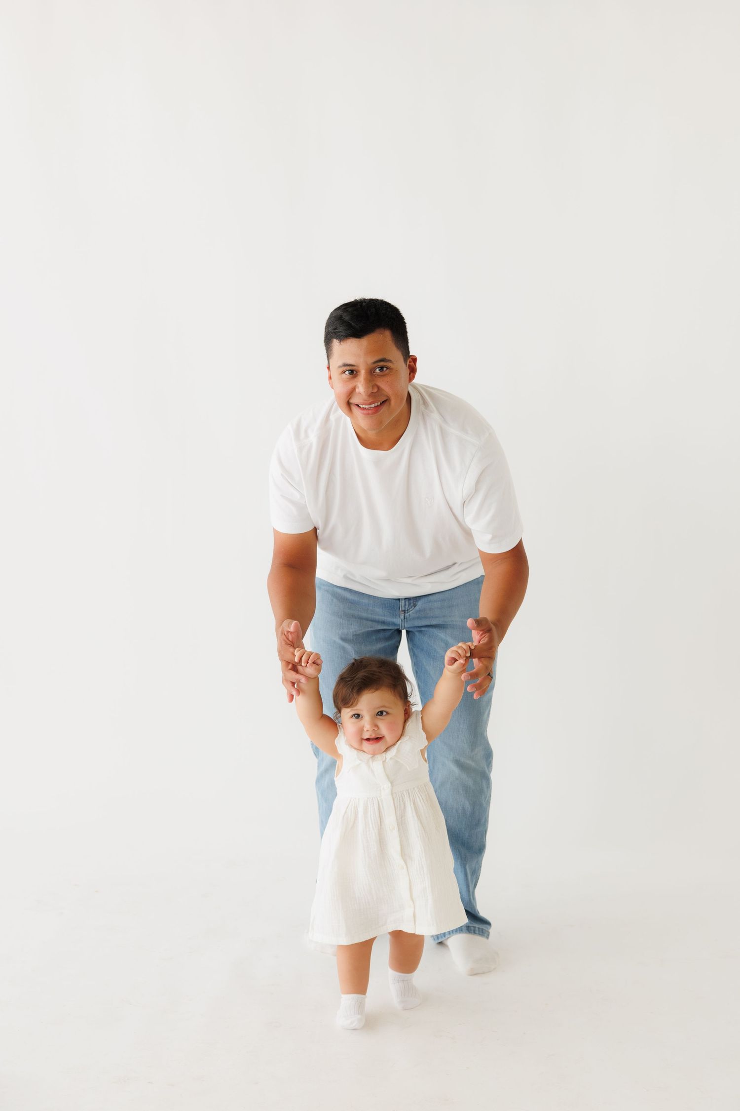 Father helping toddler daughter take first steps on white background.