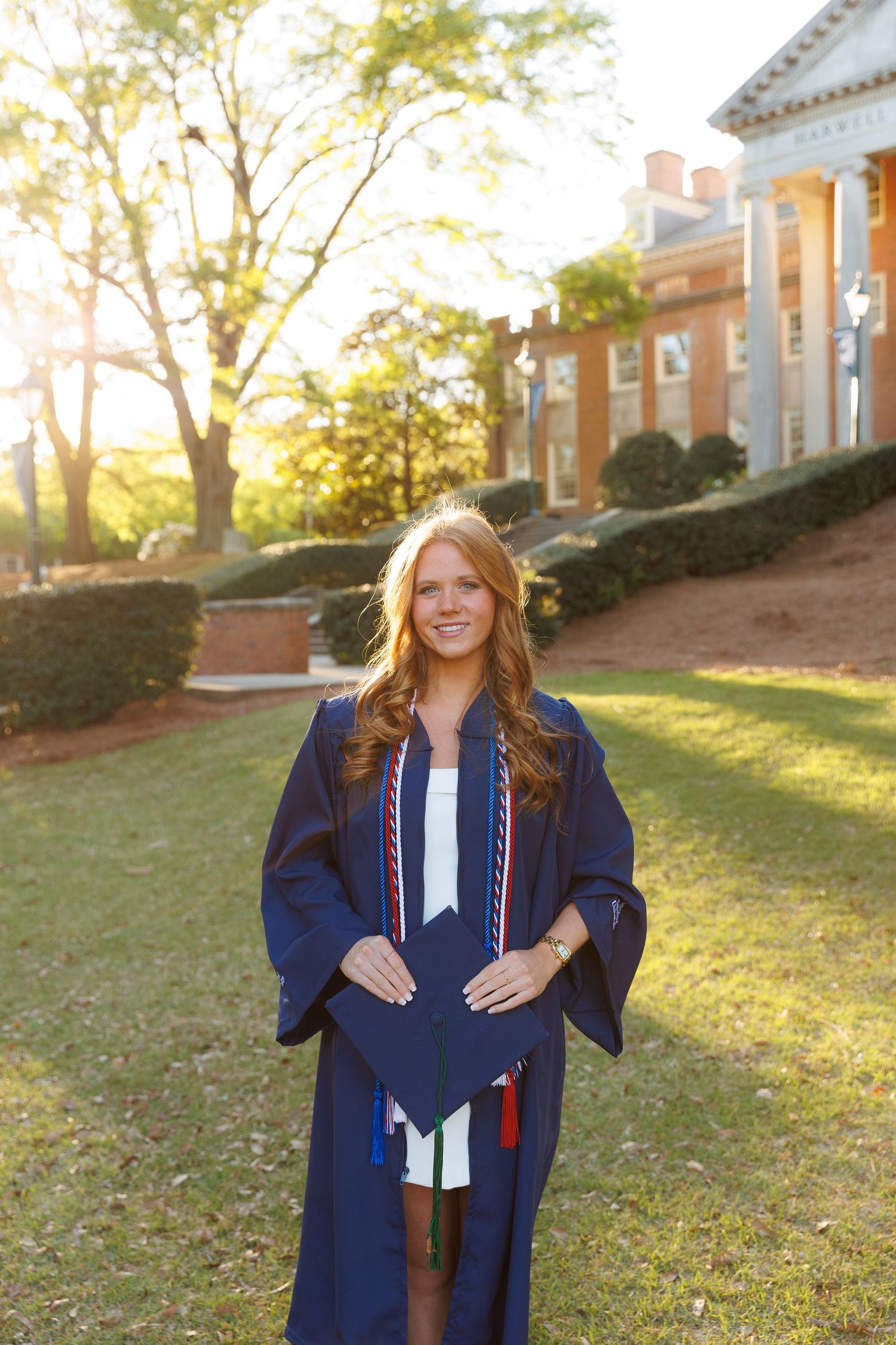Young woman in navy blue graduation gown with honor cords smiling outdoors on a college campus.