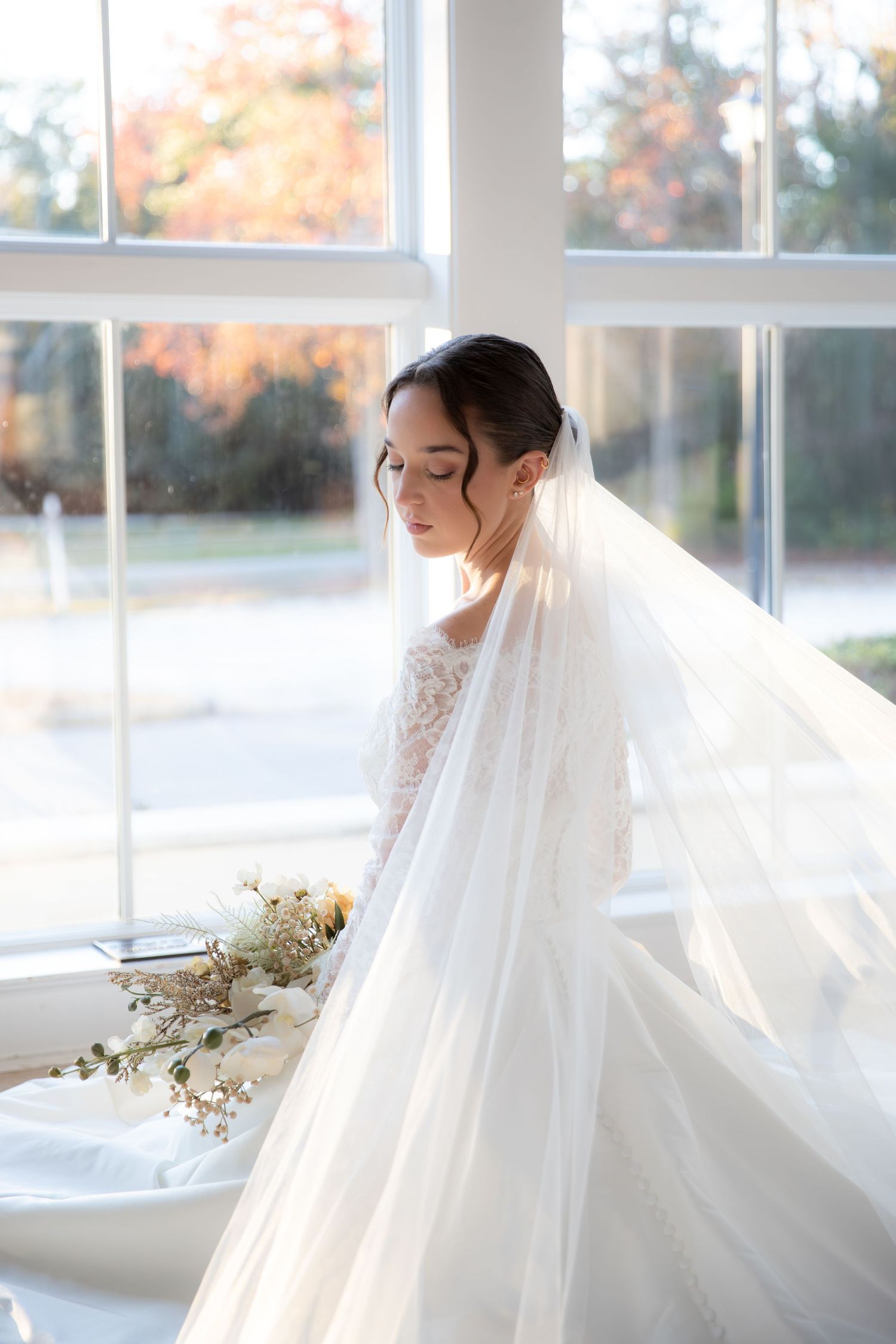 A flowing white wedding veil catches the sunlight through large windows in an ethereal bridal portrait.