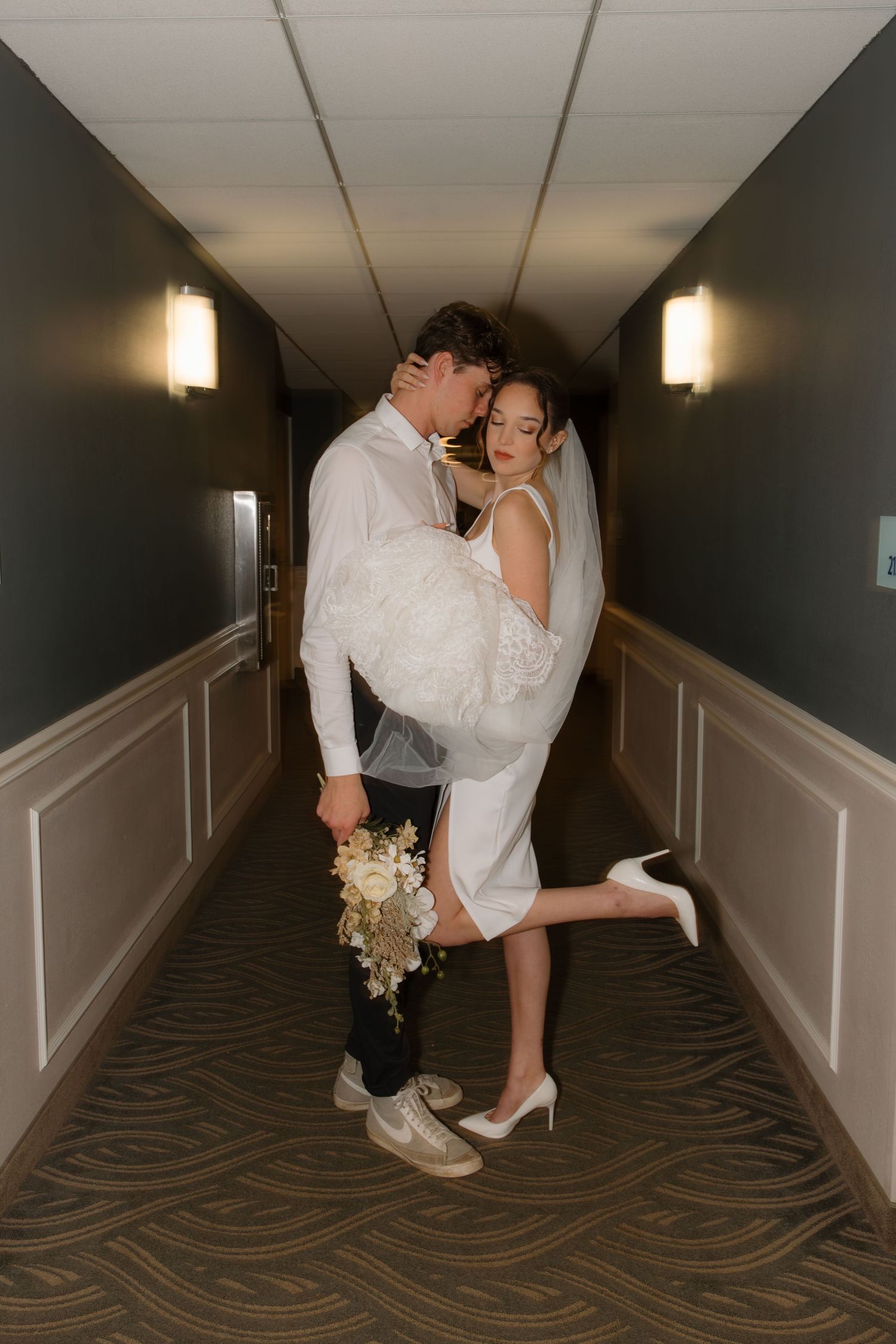 A couple shares intimate moments in a dimly lit hotel hallway with decorative carpet and wall sconces.