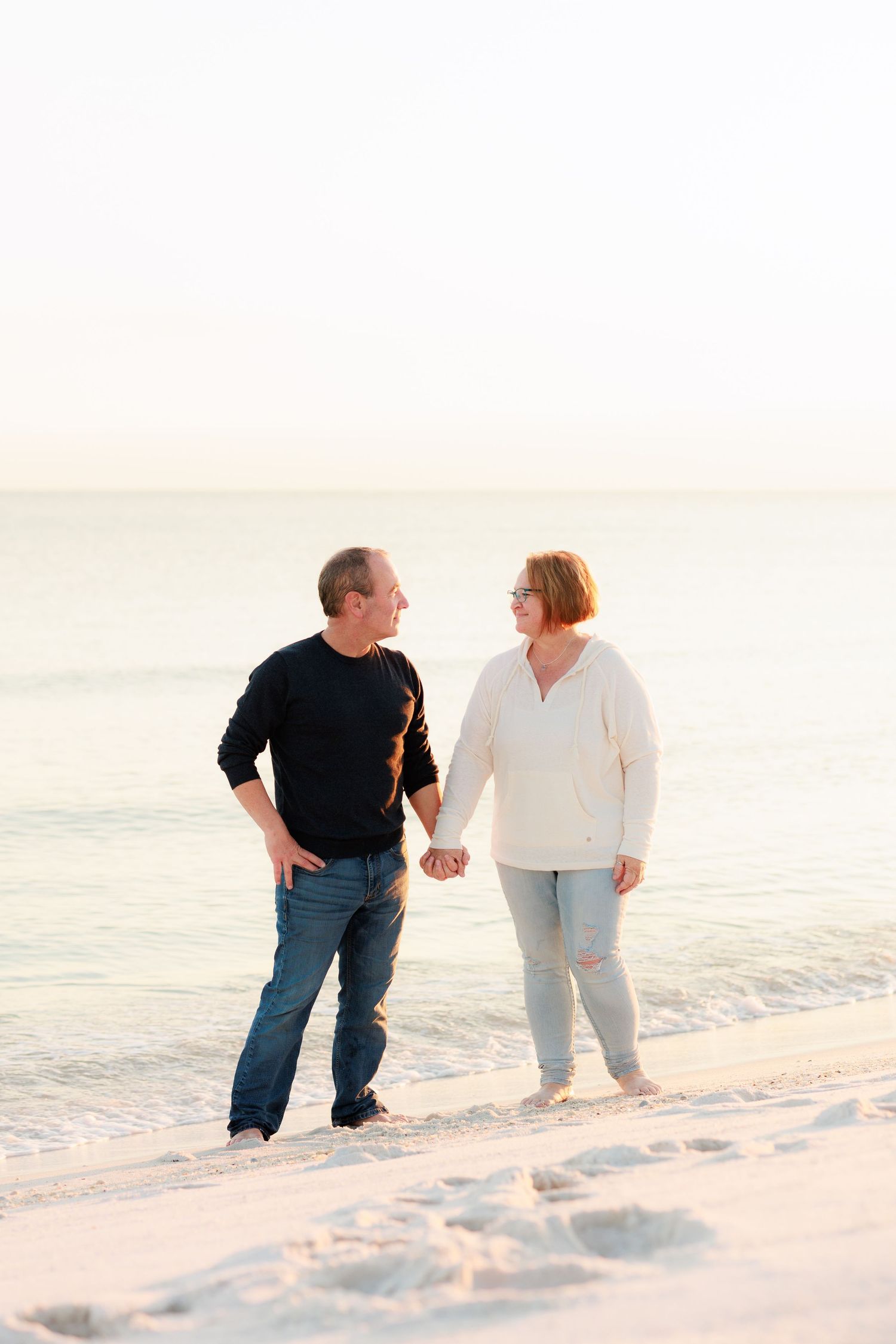 A couple walking together hand in hand on a white sandy beach at sunset in casual attire.