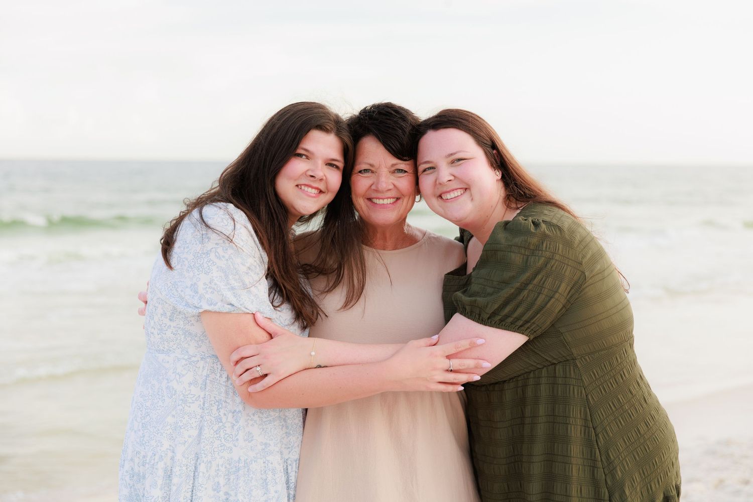 Three people embracing and smiling together on a sunny beach with ocean waves visible in the distance.