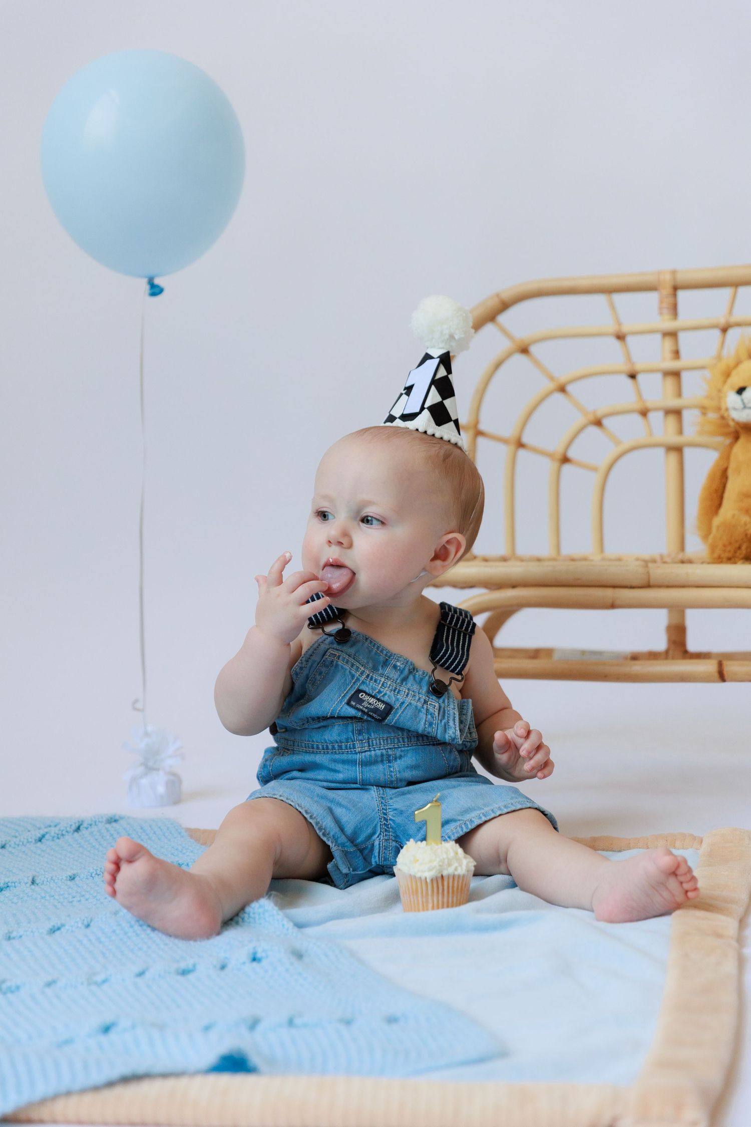 Baby in denim overalls sits on blue blanket with birthday cupcake and balloon for first birthday celebration.