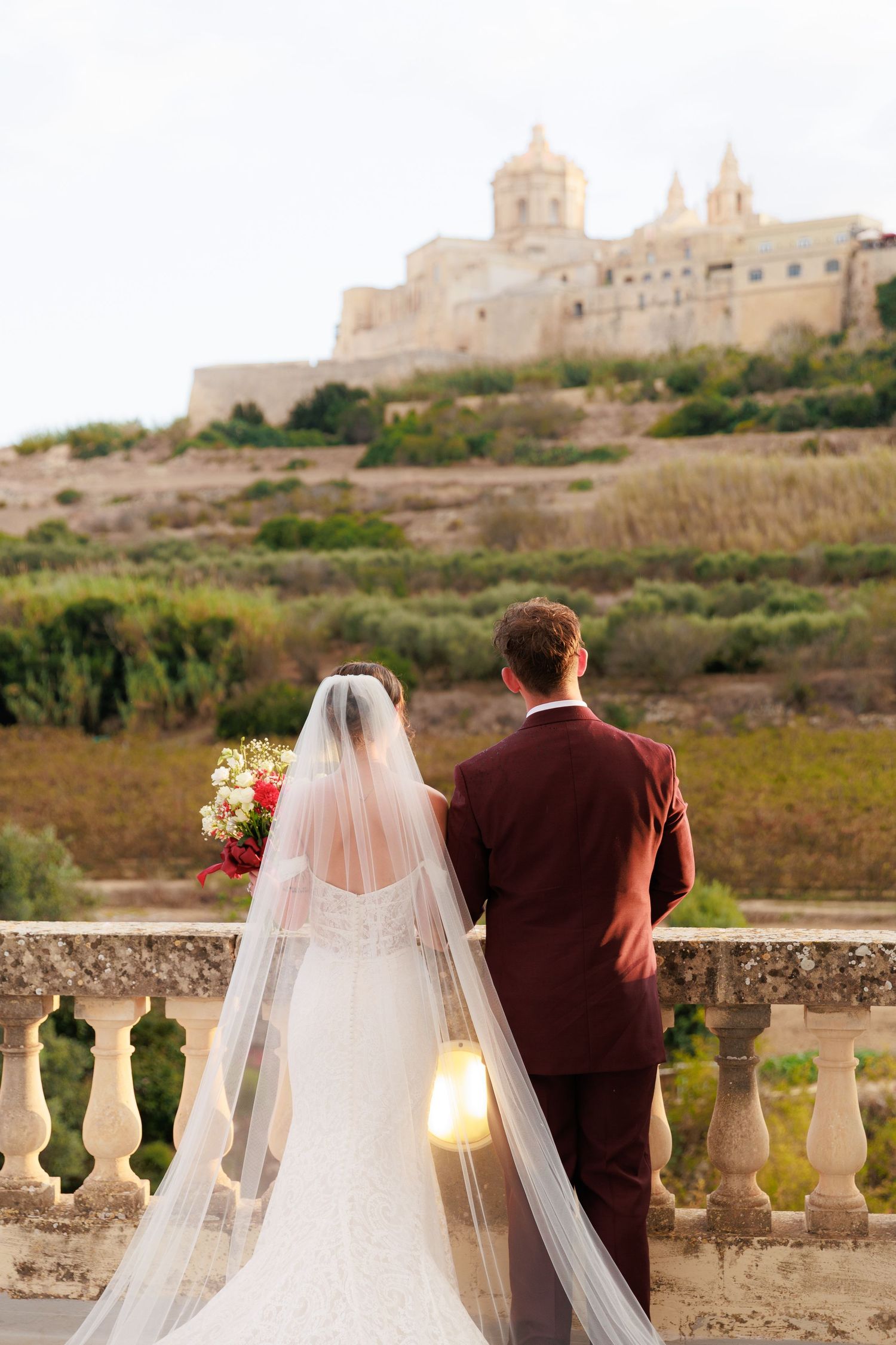 A couple stands at a stone balustrade overlooking a scenic Mediterranean landscape with historic buildings in the distance.