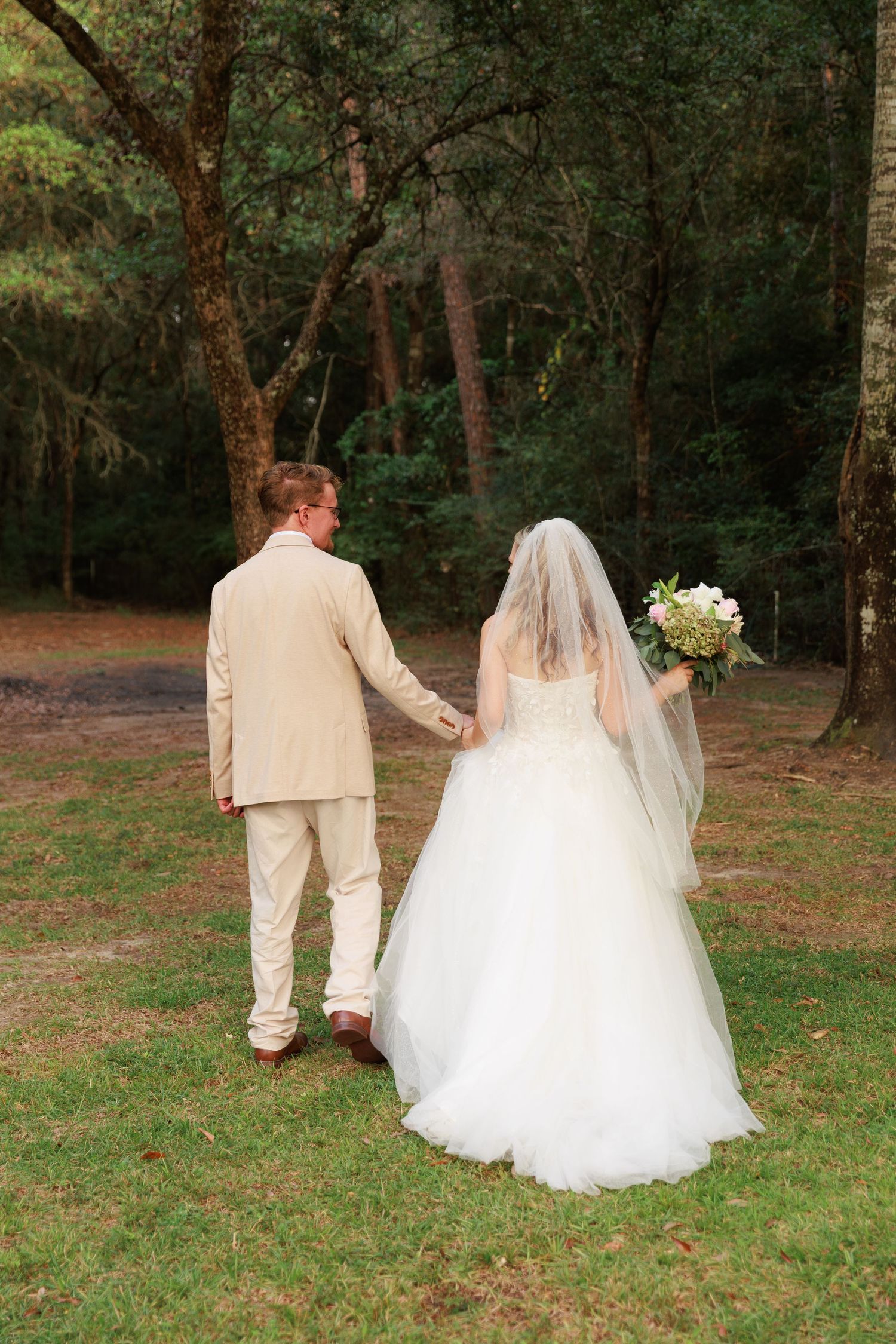 A couple in wedding attire walks hand in hand through a wooded outdoor setting.