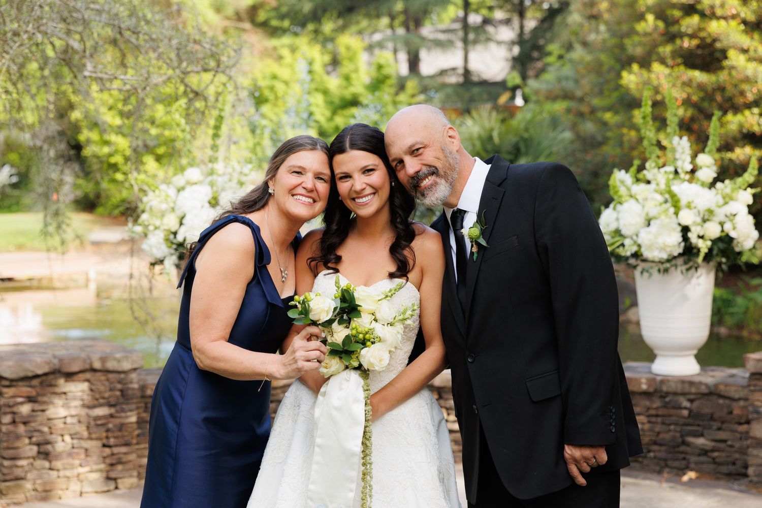 Bride in white gown poses with bridesmaid in navy dress and bald man in black suit at outdoor wedding ceremony.