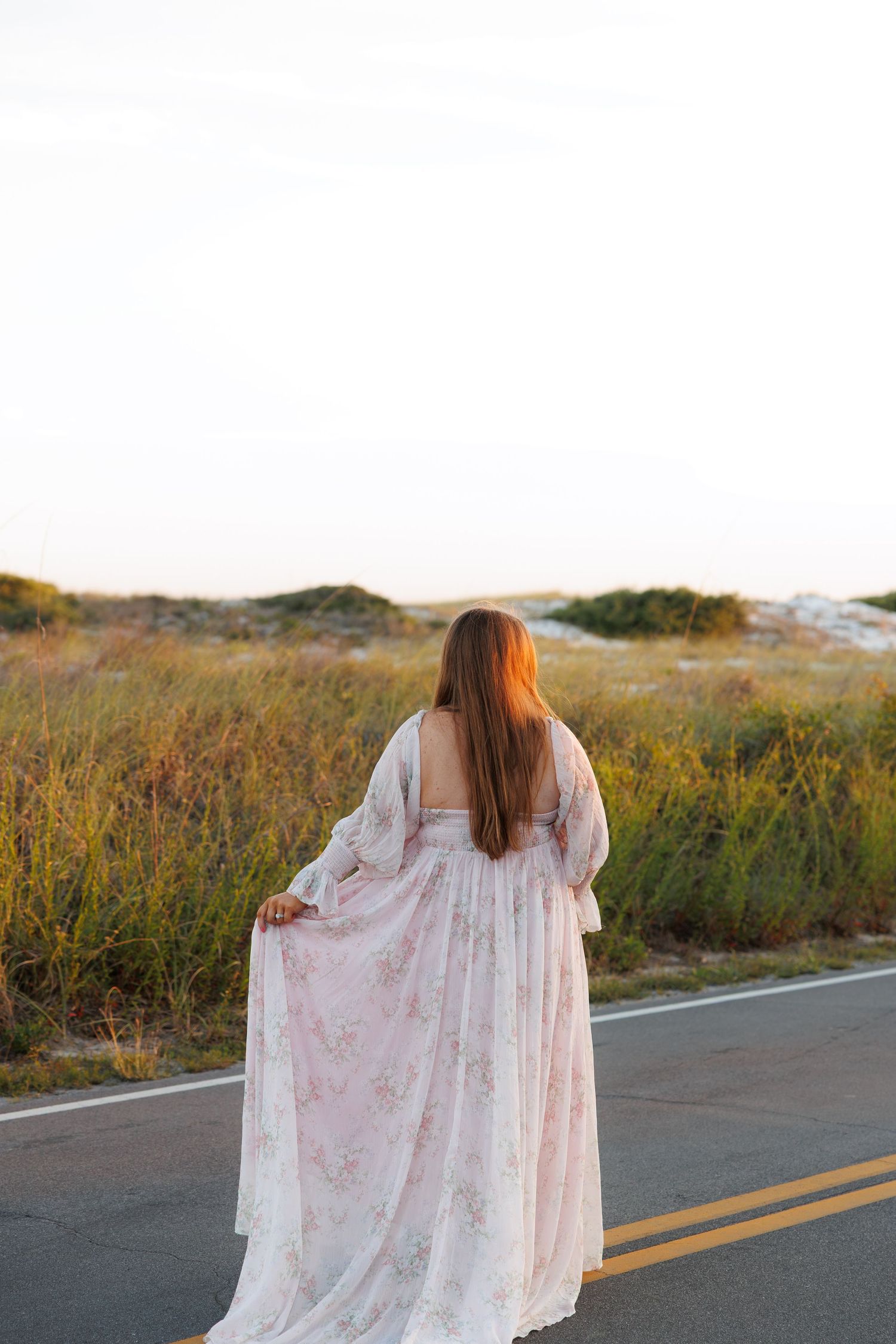 Pregnant woman in flowing floral maternity gown walks along coastal road at golden sunset.
