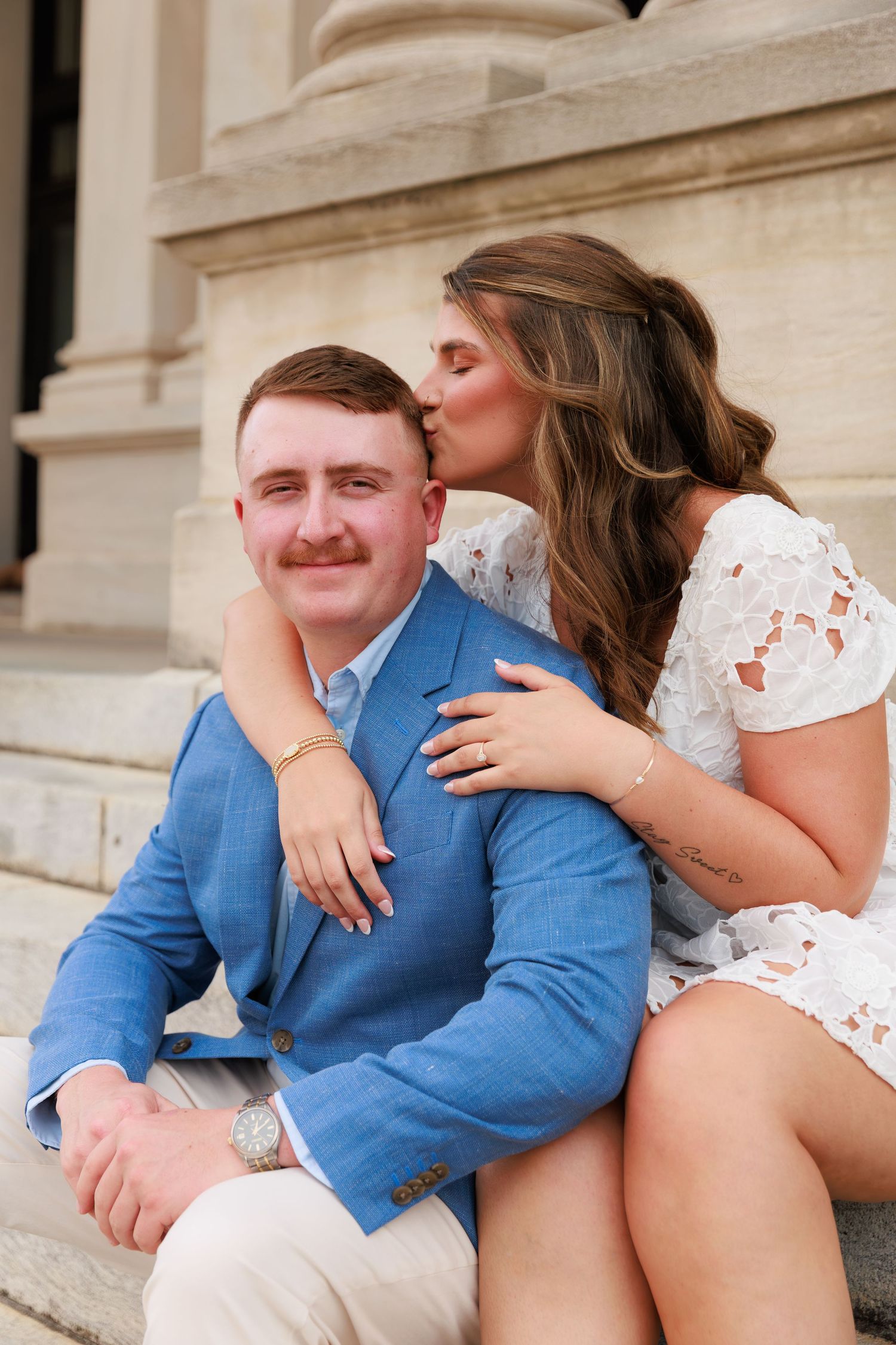 Woman in white lace dress kisses man in blue blazer on the cheek while sitting on stone steps.