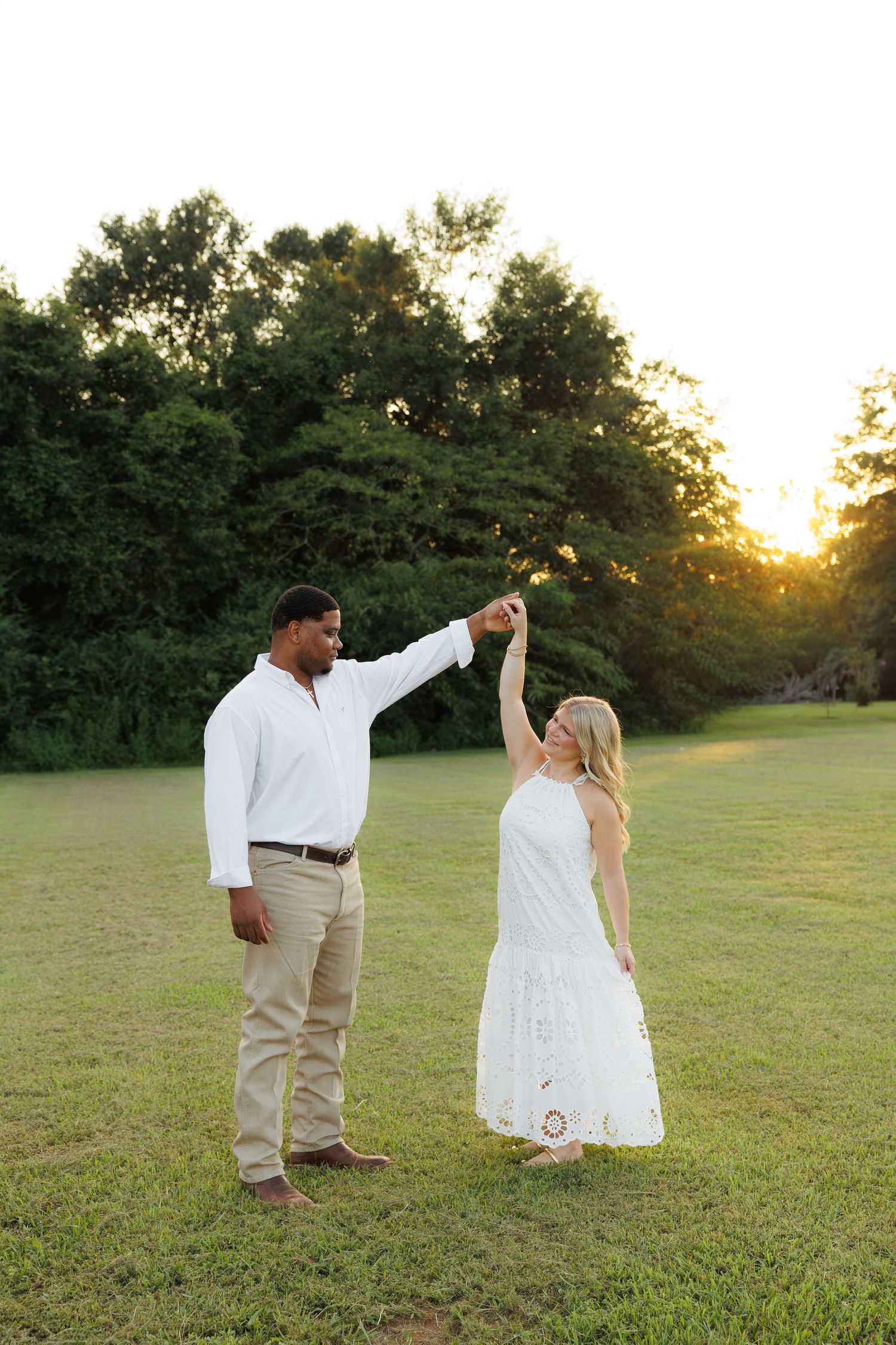 Couple dancing at sunset in a green field, man in white shirt twirling woman in white lace dress.