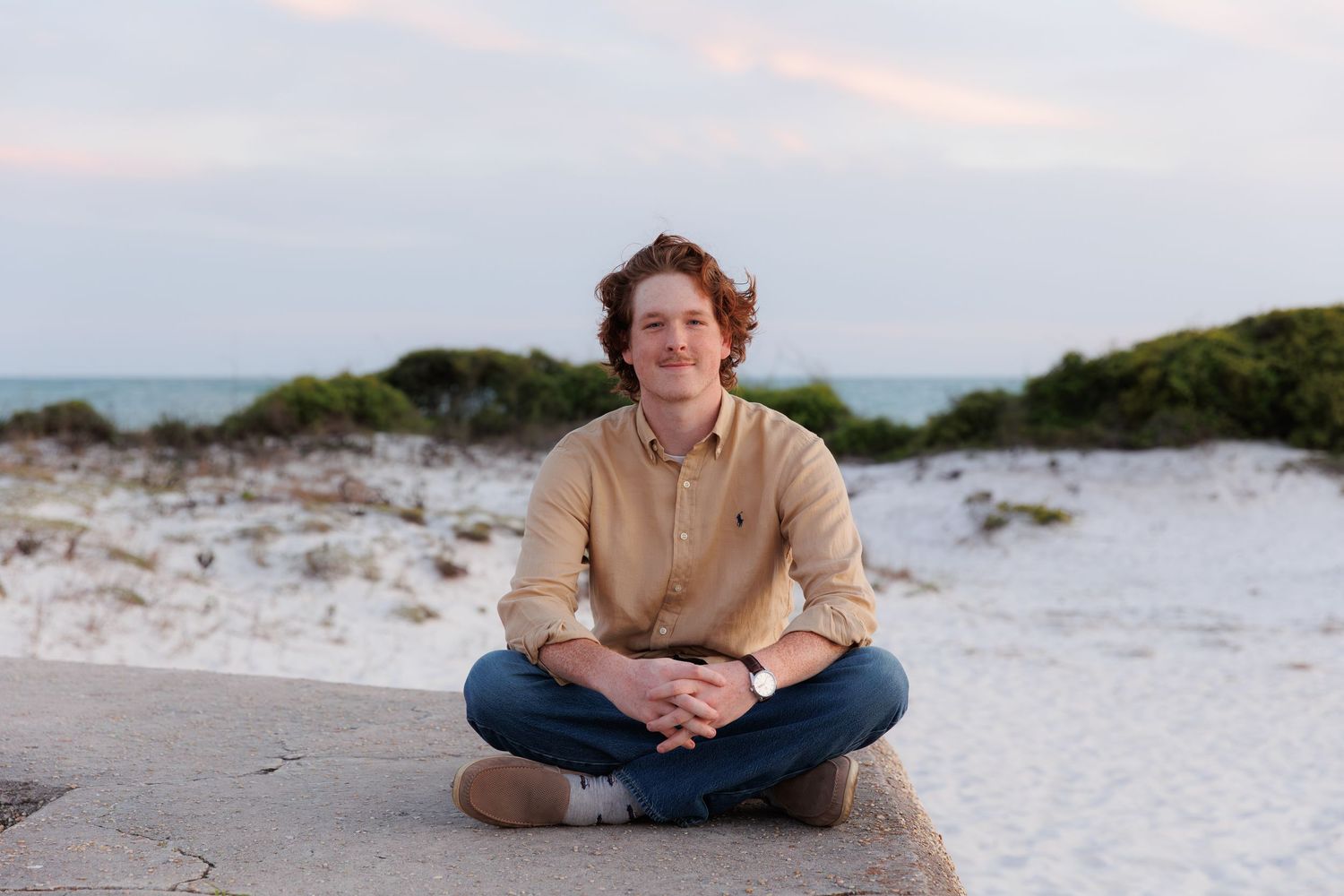 Young man with curly red hair sits cross-legged on a beach wall, smiling, wearing a tan shirt and jeans.