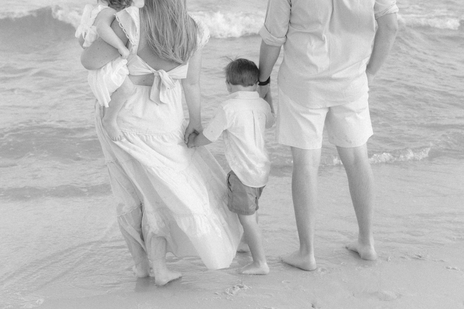 Black and white beach photo of a family in white outfits, parents holding children near ocean waves.