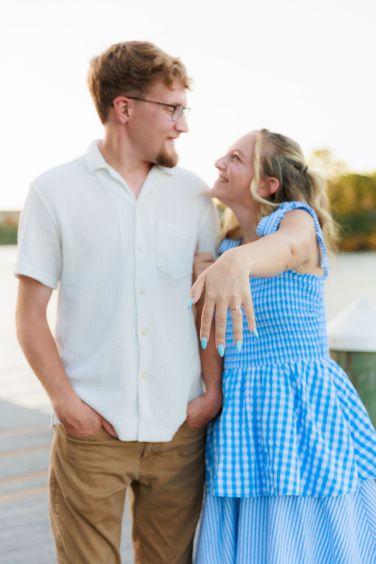 A young couple smiles at each other on a dock, woman in blue gingham dress hugging man in white shirt.