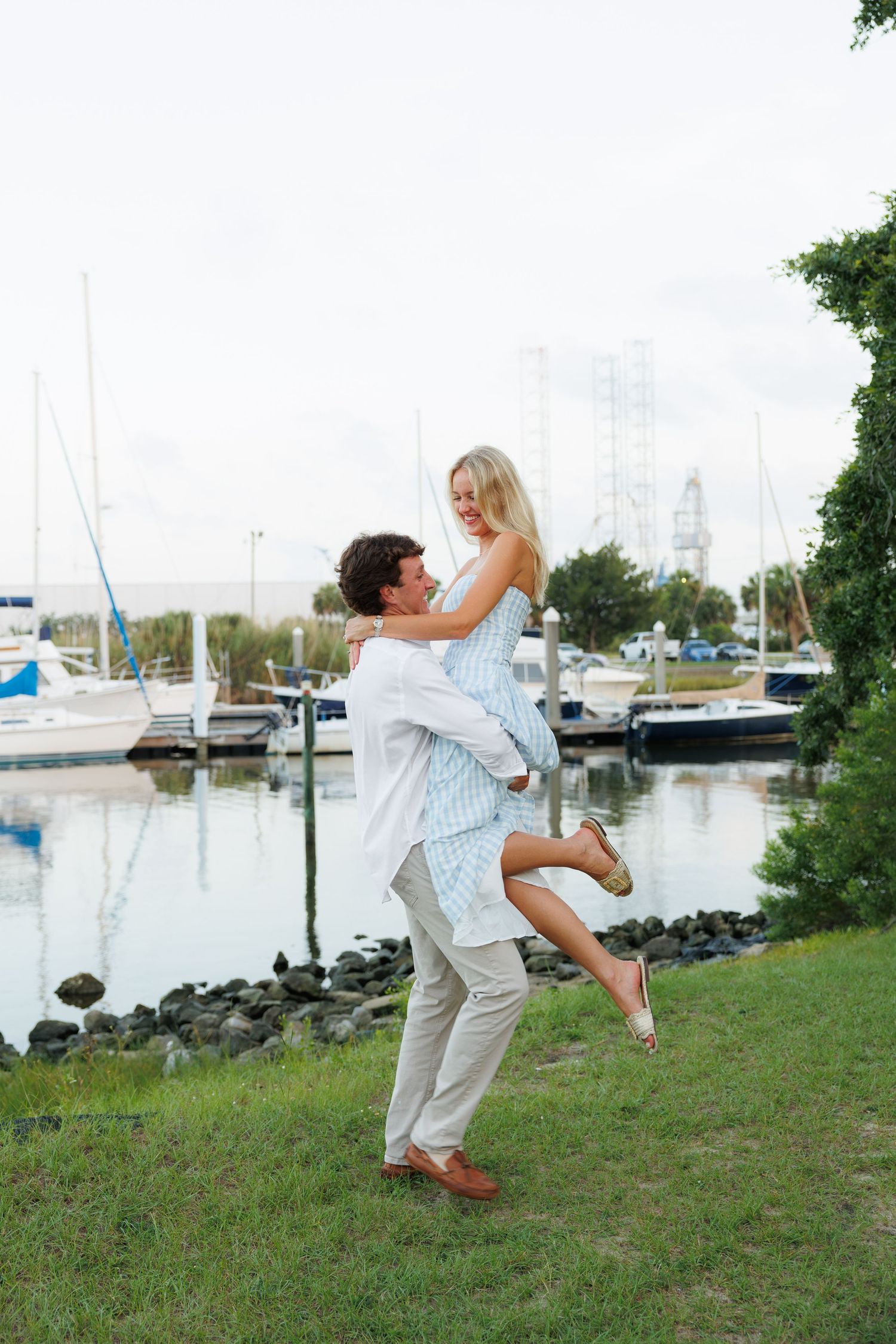 A man lifts a smiling blonde woman near a marina with sailboats and calm water in the background.