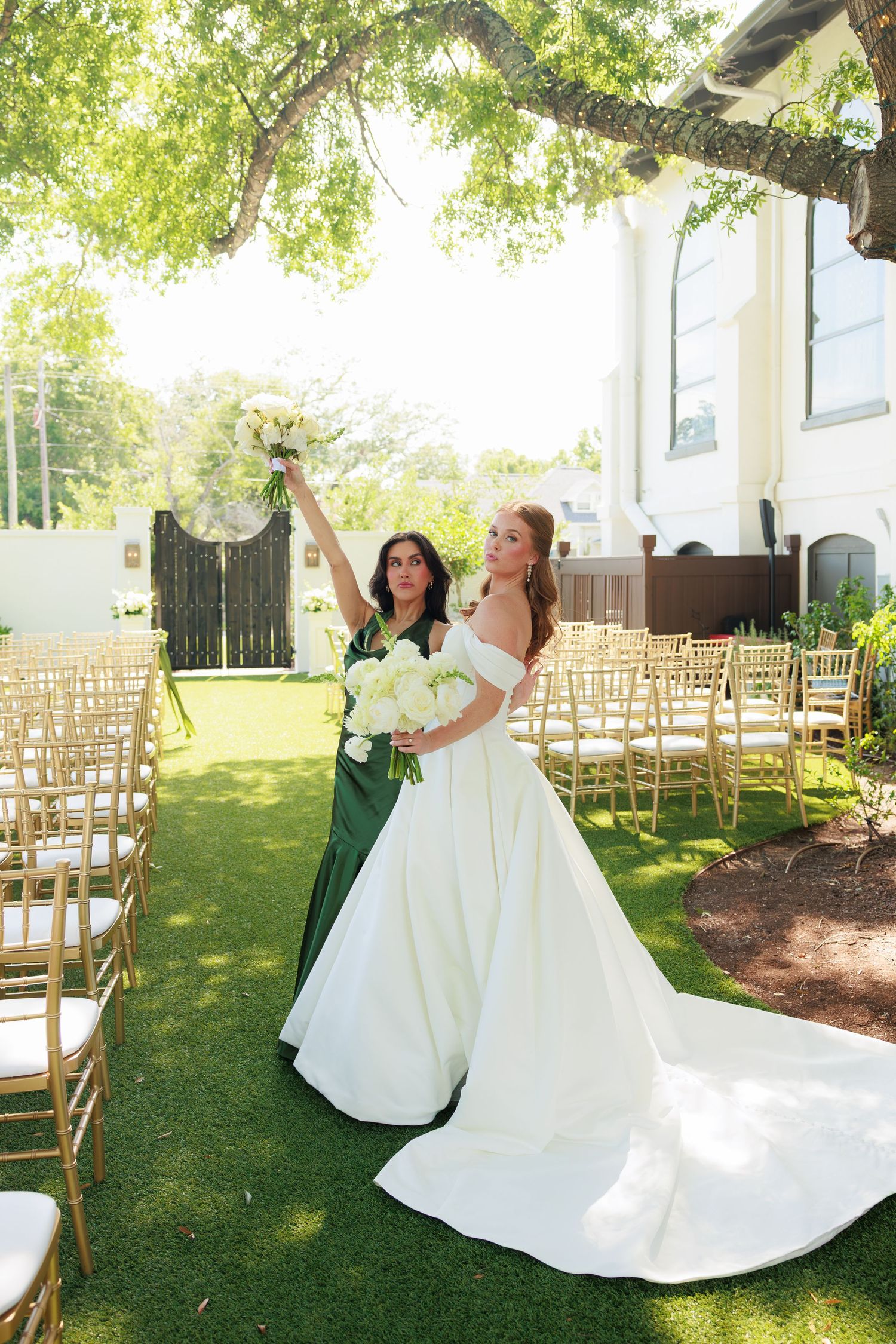 Bride in white gown and bridesmaid in green dress celebrate outdoors at elegant garden wedding ceremony with gold chairs.